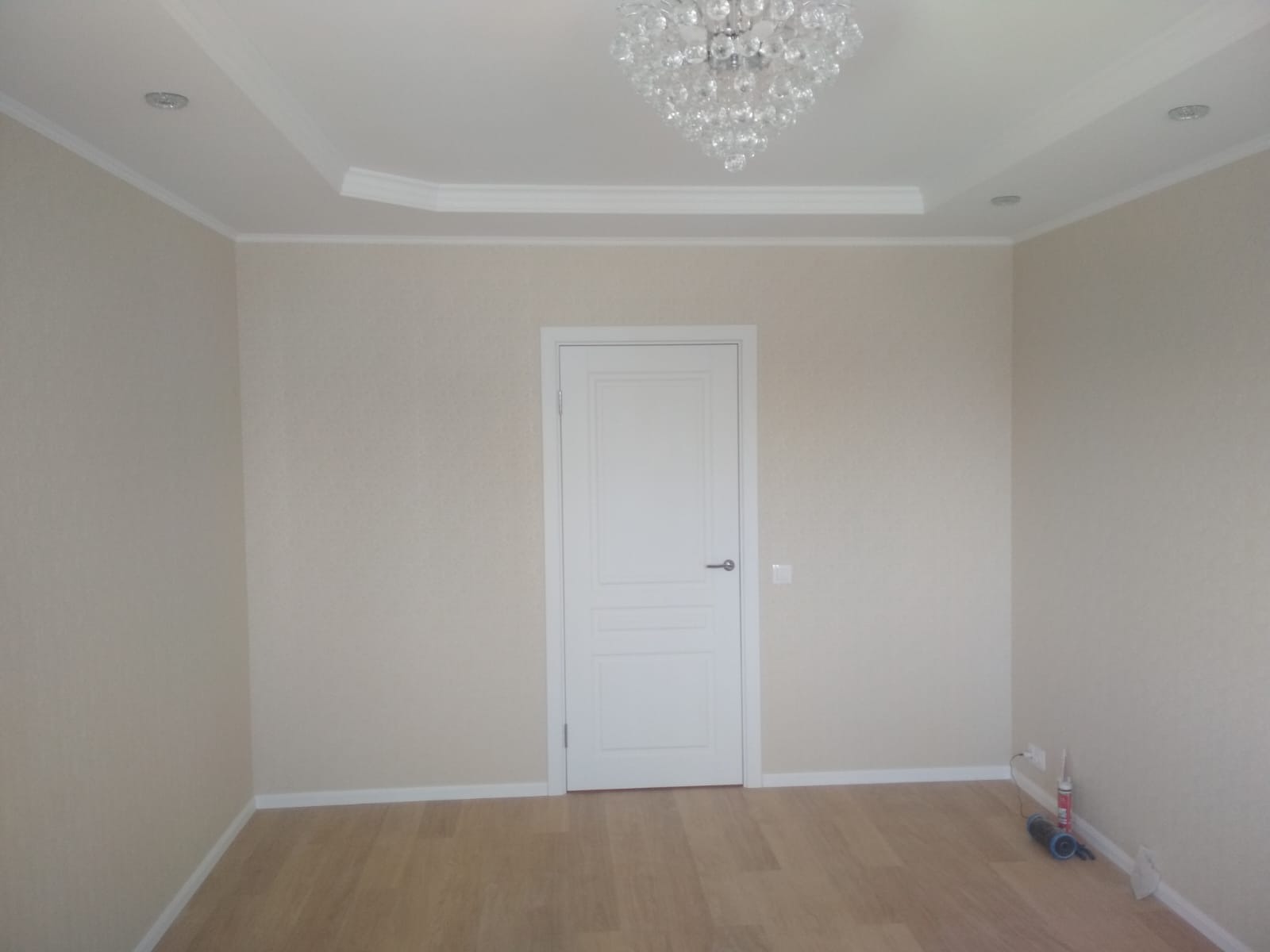 Jasne wnętrze po remoncie: kremowe ściany, drewnopodobna podłoga, białe drzwi i listwy przypodłogowe. Kryształowy żyrandol i gładki sufit z oświetleniem punktowym.