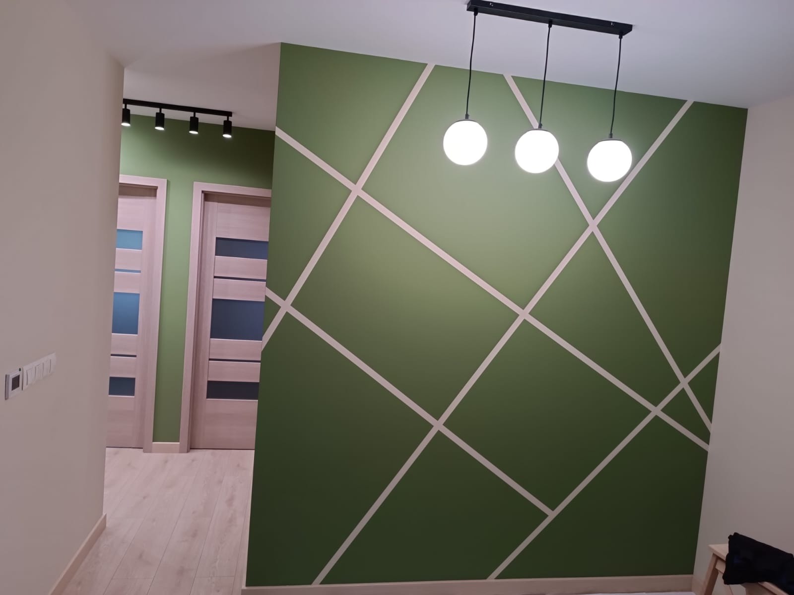 Zielona ściana z geometrycznym wzorem z jasnych pasów, oświetlona trzema kulistymi lampami. W tle widoczne drzwi i oświetlenie szynowe.