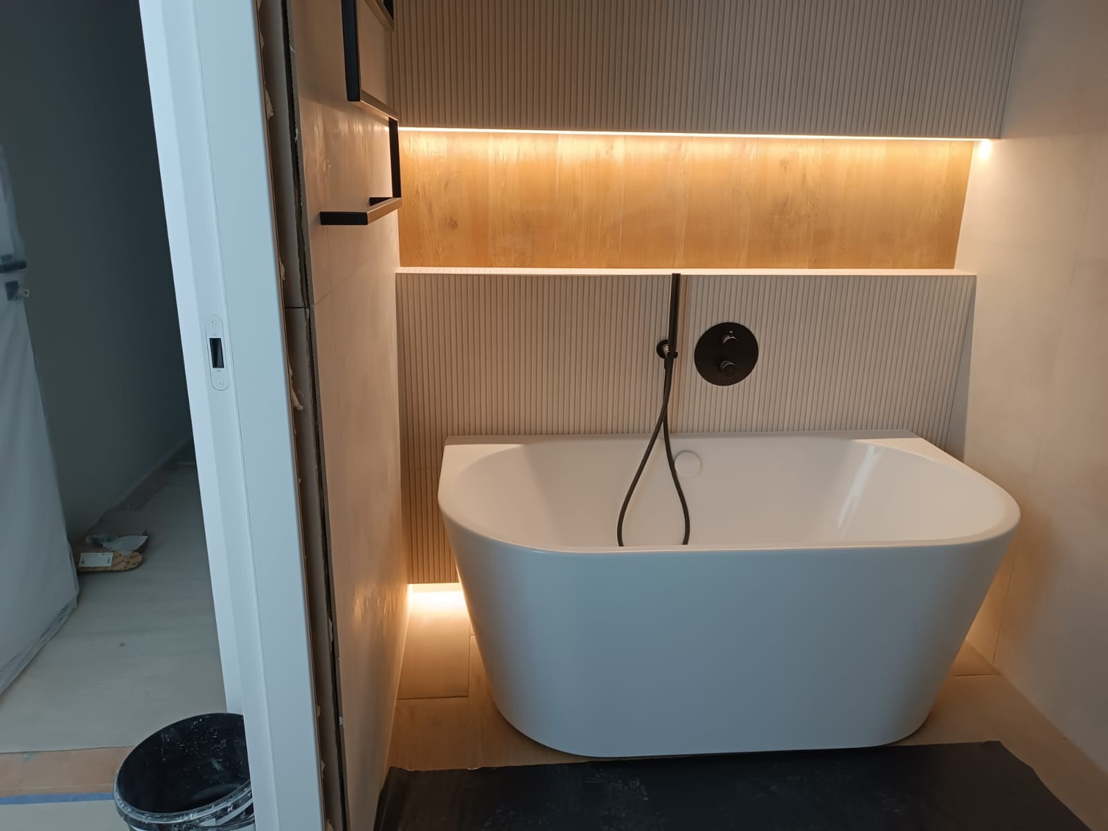 Nowoczesna, wolnostojąca wanna z czarną armaturą na tle ściany z podświetlanymi półkami. Widoczny fragment otwartych drzwi i wiadro. Minimalistyczny design łazienki.