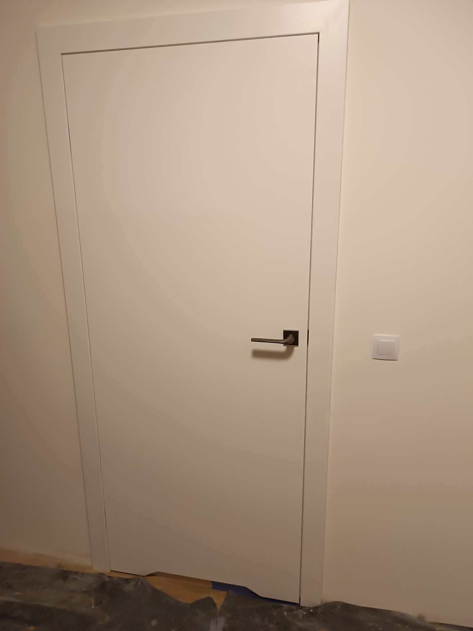 Białe, minimalistyczne drzwi wewnętrzne z ciemną klamką na tle jasnej ściany i fragmentu podłogi zabezpieczonej folią malarską. Widoczny włącznik światła.