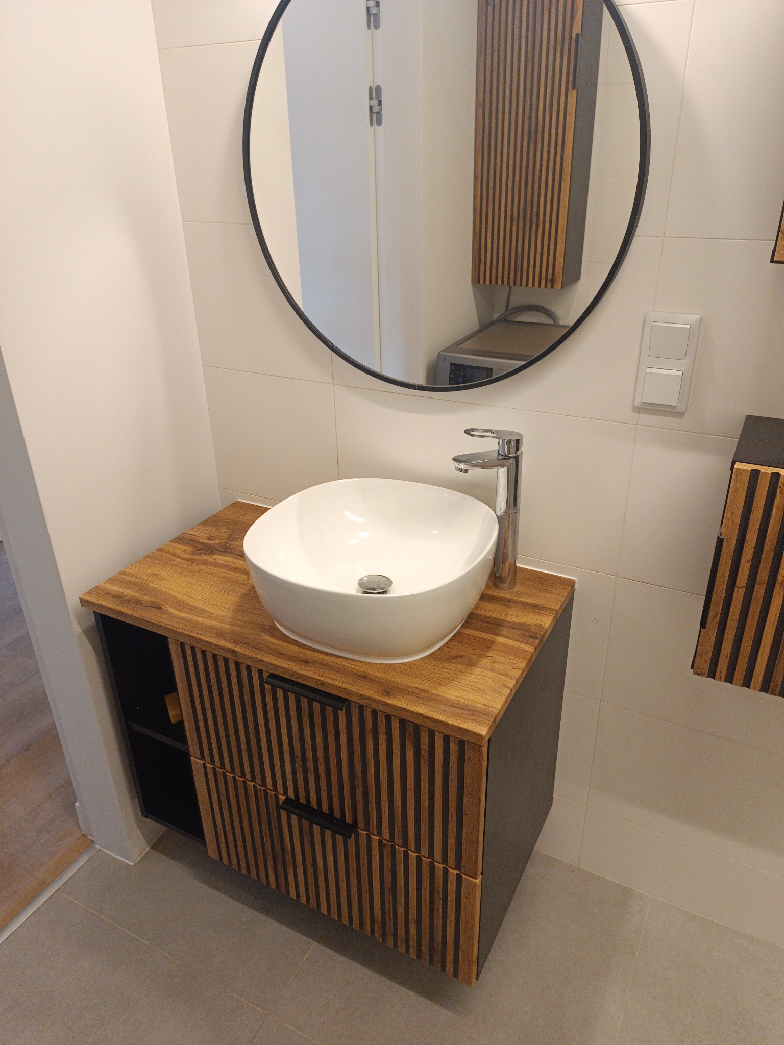Nowoczesna łazienka z drewnianą szafką pod umywalkę i okrągłym lustrem. Umywalka biała, nablatowa. Widoczne płytki na ścianach i podłodze w neutralnych barwach.