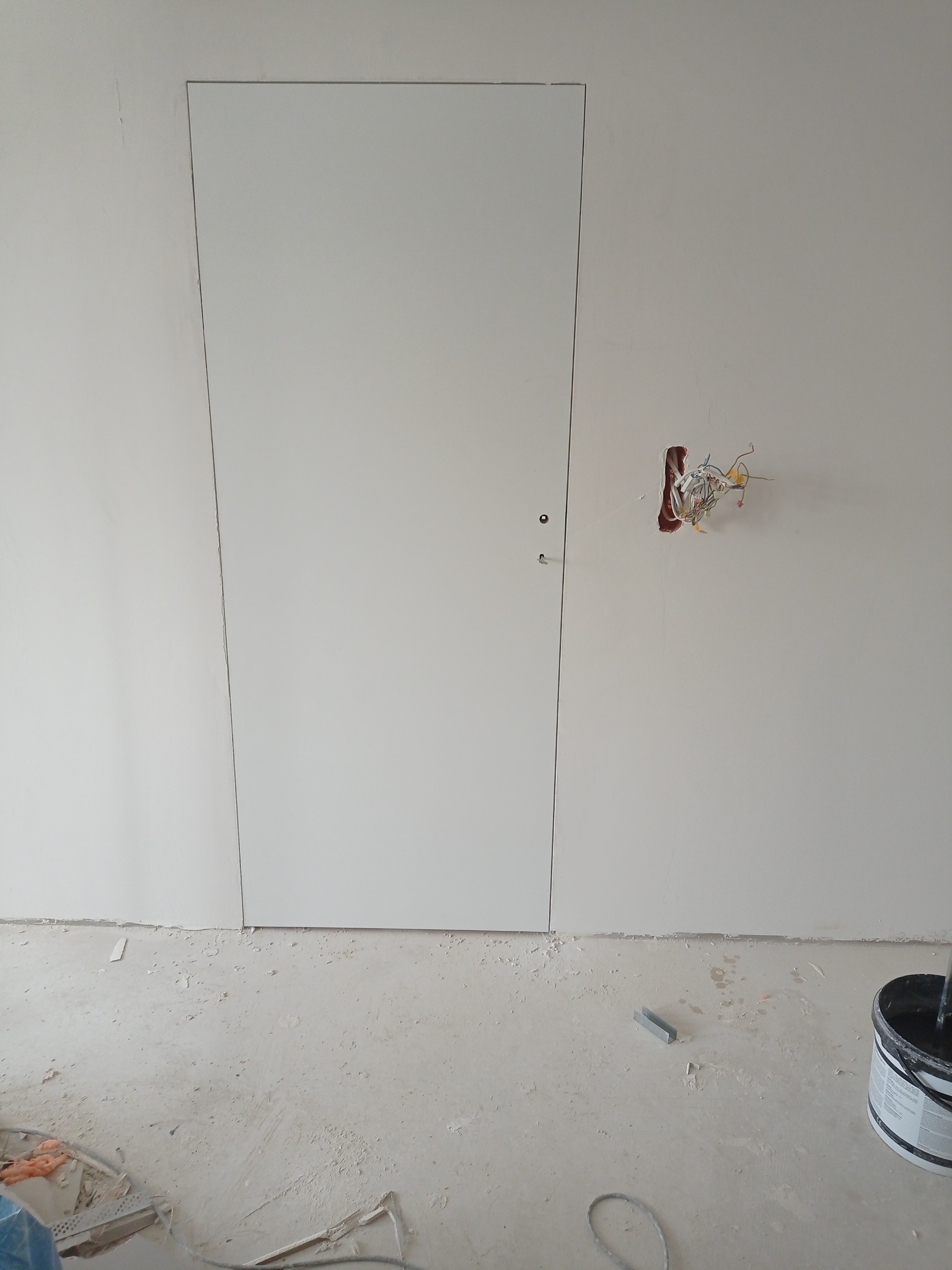 Białe, ukryte drzwi w ścianie po szpachlowaniu, z widoczną puszką elektryczną i przewodami. Podłoga w trakcie prac wykończeniowych.