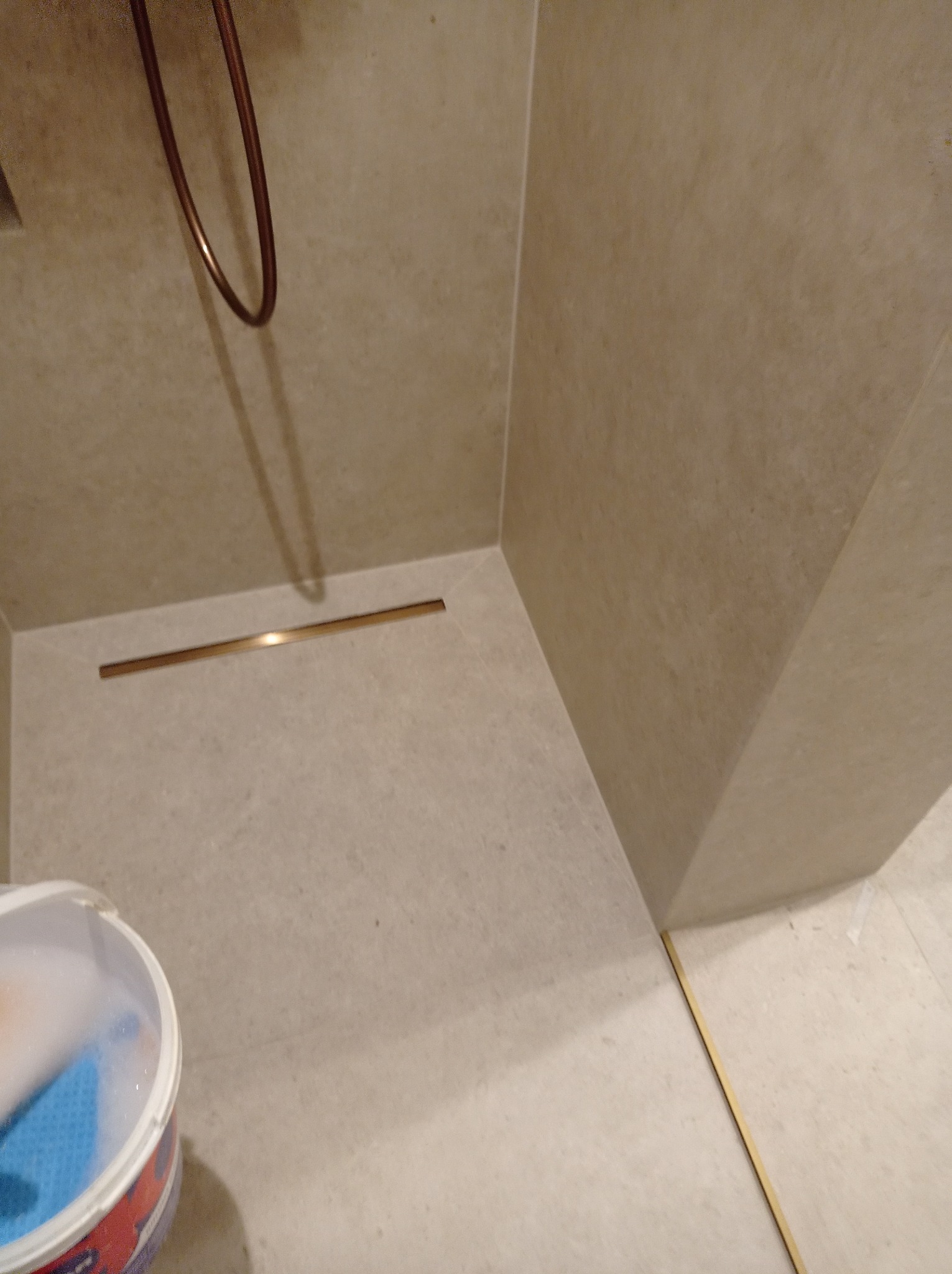 Beżowa kabina prysznicowa z odpływem liniowym w kolorze złotym. Widoczny fragment baterii i wiadro z akcesoriami do czyszczenia. Minimalistyczny design.