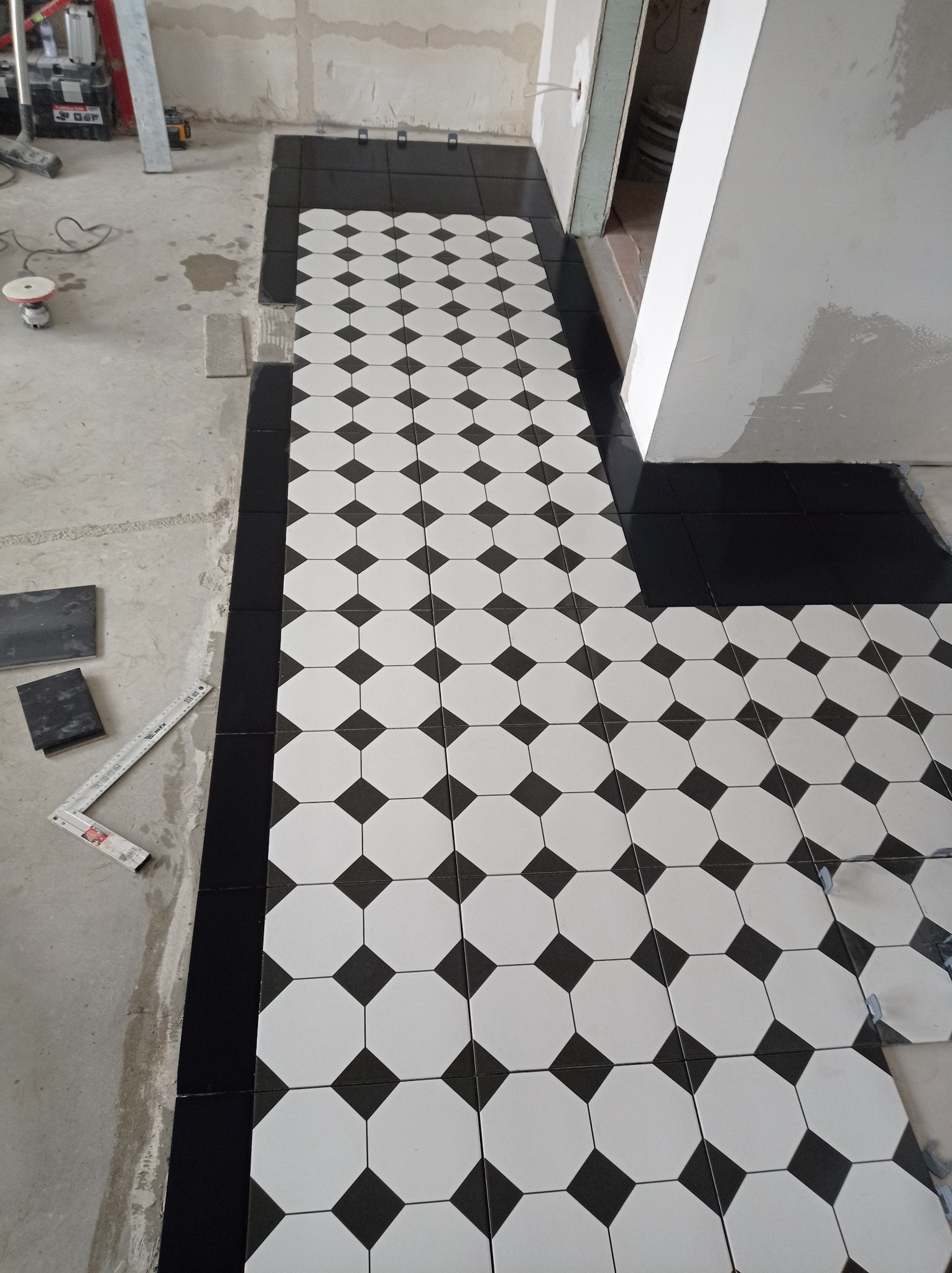 Układanie czarno-białej mozaiki podłogowej w romby, z czarną obwódką. Widoczne narzędzia glazurnicze i surowy beton. Praca w toku, fragment podłogi wykończony.