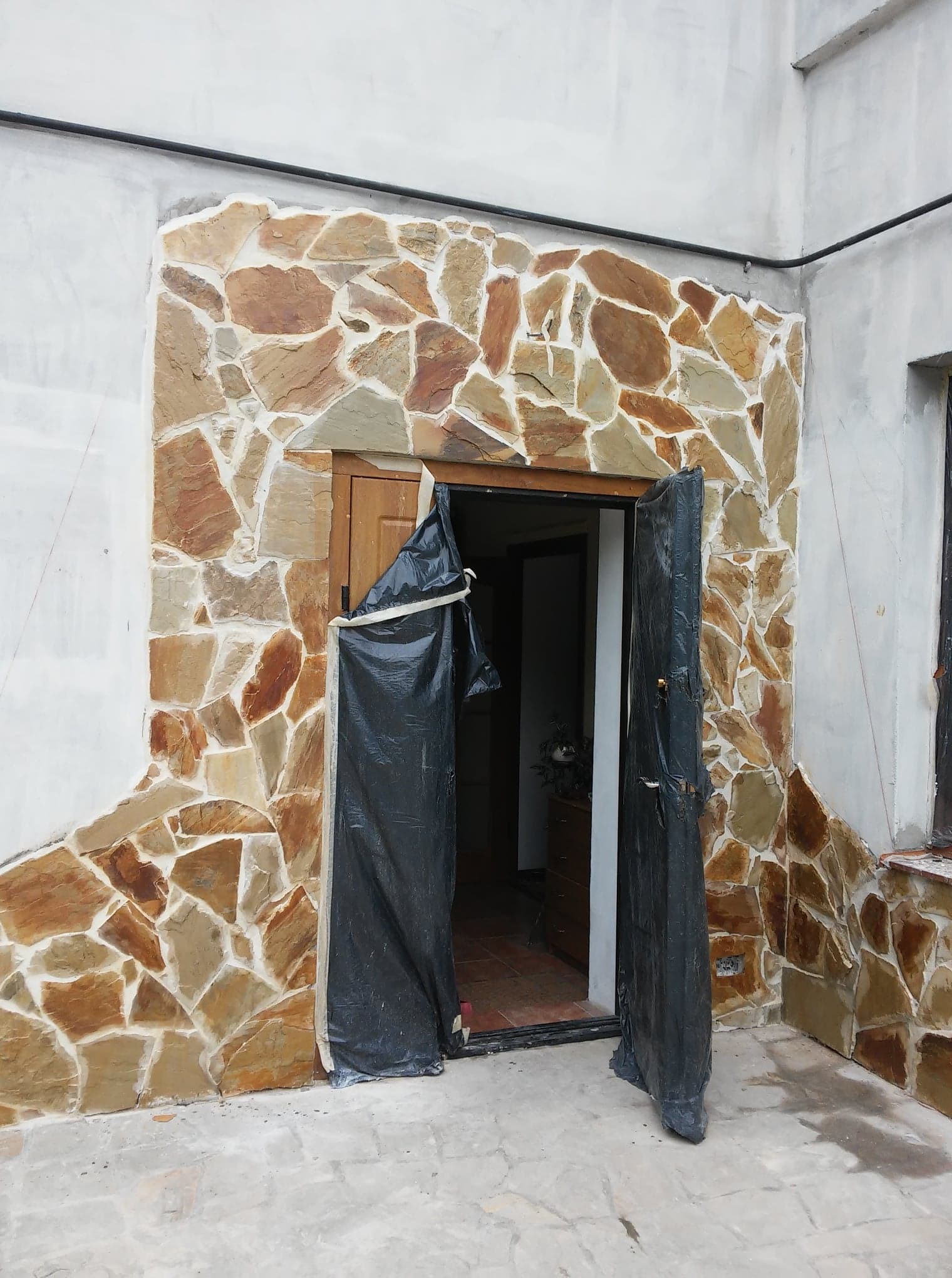 Wykończenie elewacji kamieniem dekoracyjnym wokół drzwi wejściowych, zabezpieczonych folią malarską. Widoczne fragmenty ściany z surowym tynkiem.