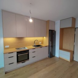 TPMU Agnieszka Rogala - Jasna kuchnia na wymiar z drewnianym blatem i czarnym zlewem, oświetlona lampą wiszącą i listwą LED, z lodówką w zabudowie i drewnianą podłogą.