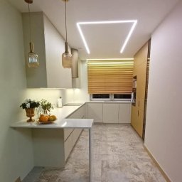 TPMU Agnieszka Rogala - Jasna kuchnia w kształcie litery U z blatem-barem, oświetleniem LED w kształcie trapezu i drewnianymi żaluzjami w oknie. Minimalistyczny design z dekoracyjnymi lampami wiszącymi.