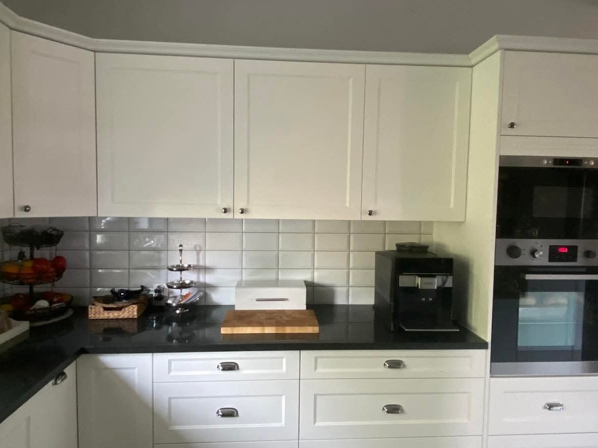 Biała zabudowa kuchenna z frezowanymi frontami, czarnym blatem i sprzętem AGD w tle. Widoczna ekspozycja z owocami i akcesoriami kuchennymi.