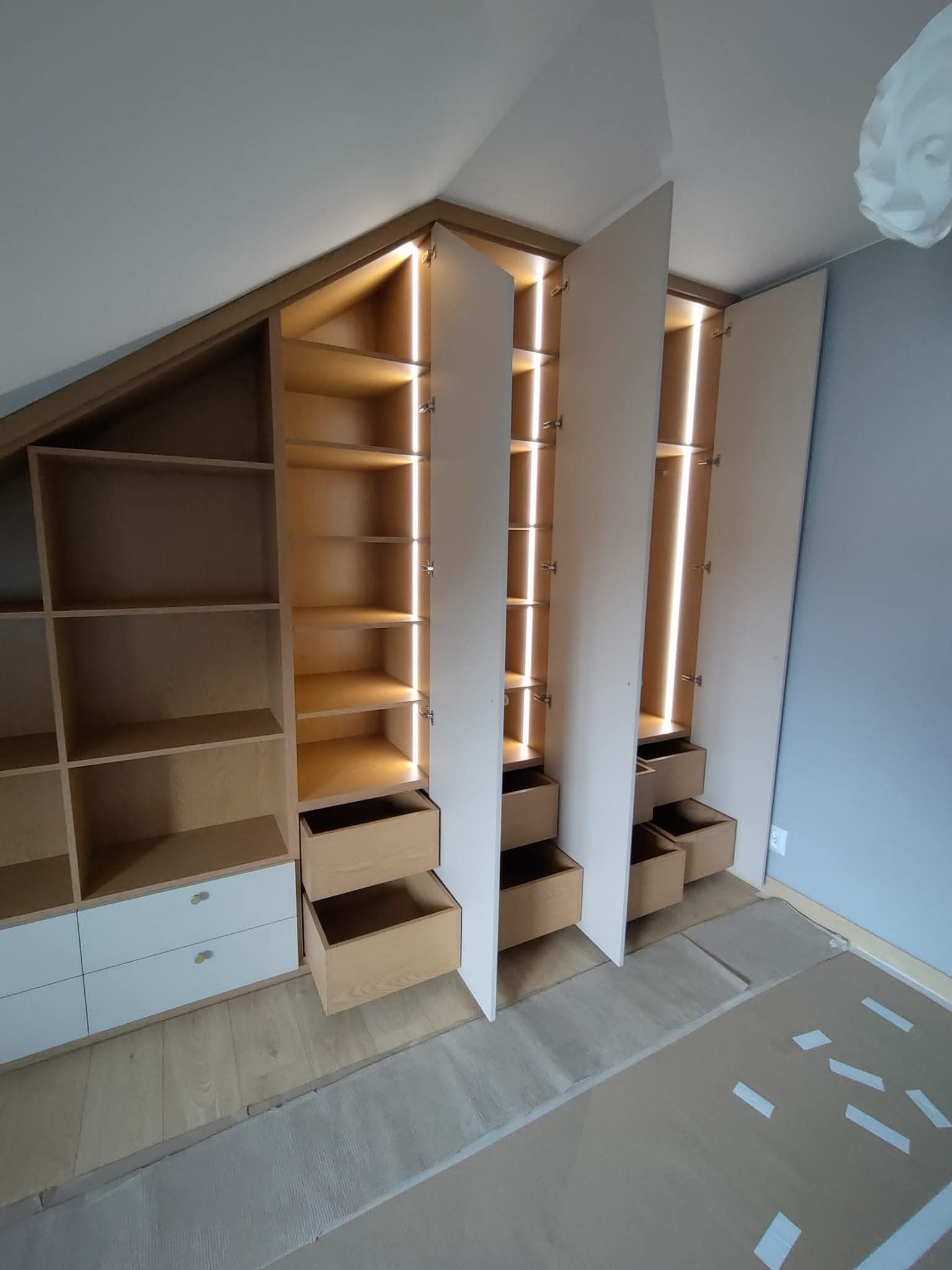 Szafa wnękowa z oświetleniem LED, jasne drewno, białe fronty, szuflady wysuwane, dopasowana do skosu dachu, widok na wnętrze szafy.