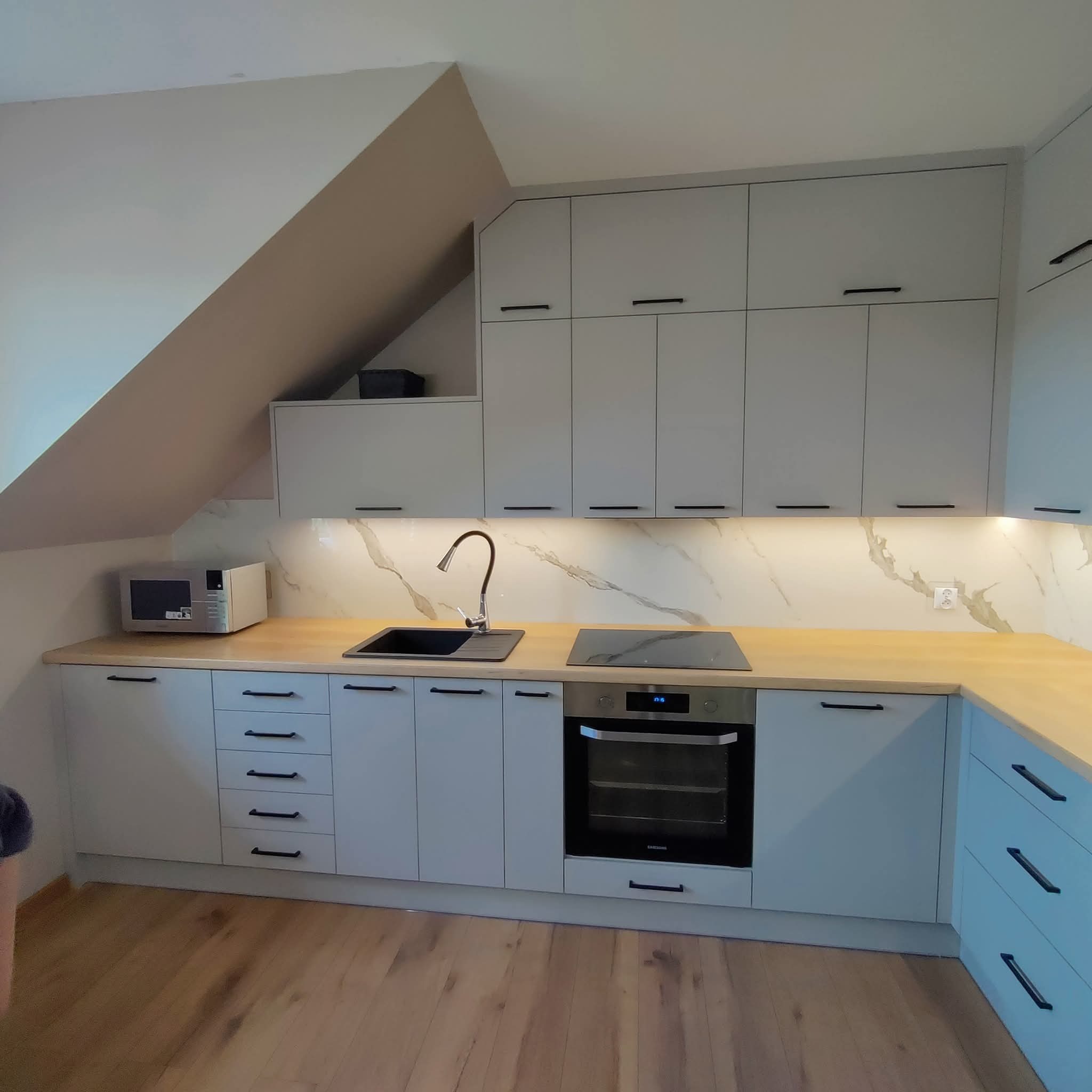 Jasna kuchnia na poddaszu z drewnianym blatem, czarnym zlewem i minimalistycznymi uchwytami. Uwagę zwraca skośny sufit i marmurowy wzór na ścianie.