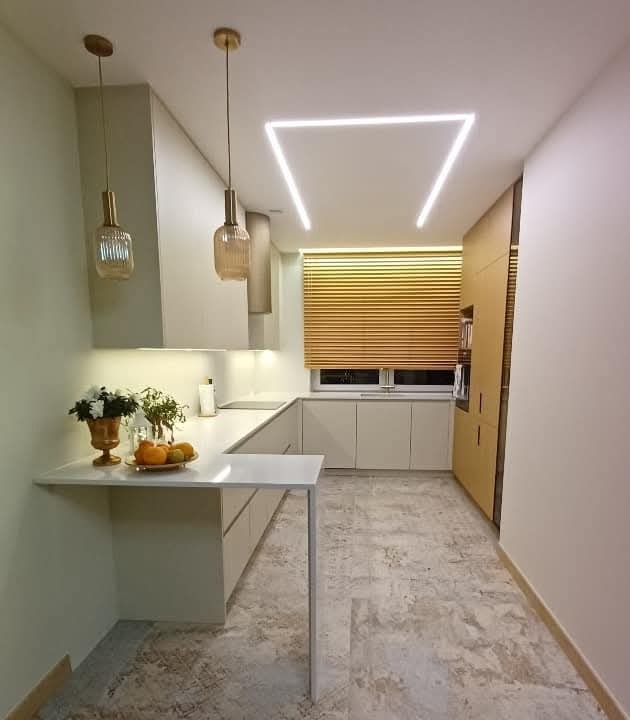 Jasna kuchnia w kształcie litery U z blatem-barem, oświetleniem LED w kształcie trapezu i drewnianymi żaluzjami w oknie. Minimalistyczny design z dekoracyjnymi lampami wiszącymi.