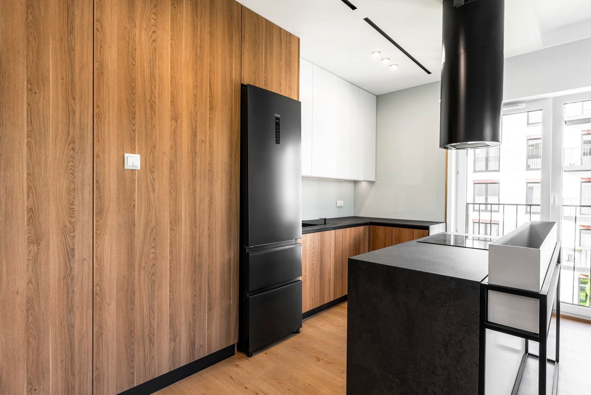 Nowoczesna kuchnia z drewnianymi frontami i czarnym sprzętem AGD w Mińsku Mazowieckim. Minimalistyczny design, wysoka zabudowa i wyspa kuchenna z czarnym blatem.