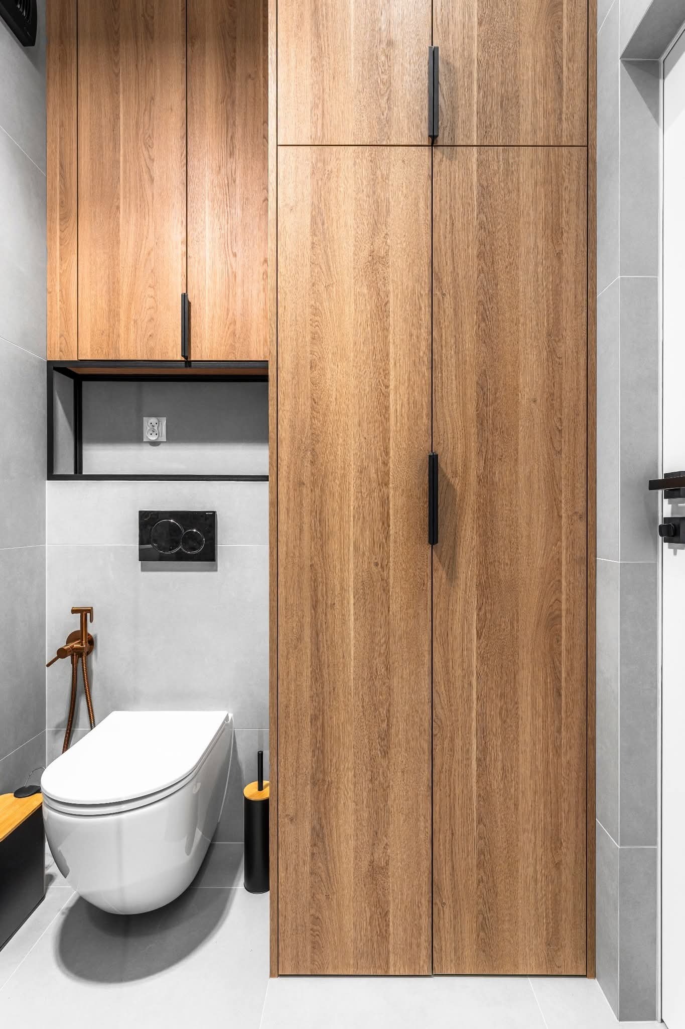 Nowoczesna łazienka z drewnianą szafą w zabudowie, wiszącą toaletą i szarą glazurą. Minimalistyczny design z akcentami czerni i naturalnego drewna.