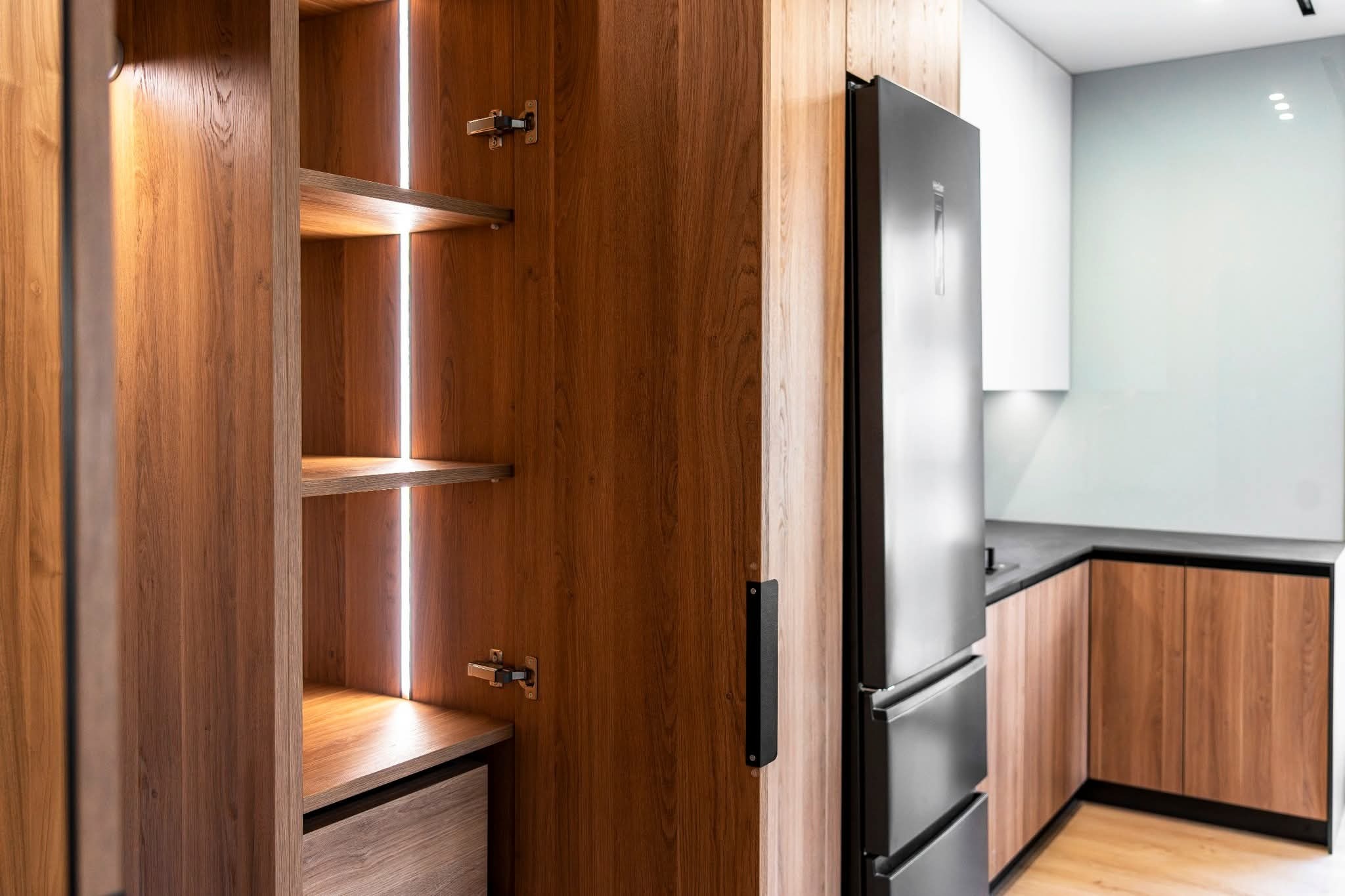 Drewniana zabudowa kuchenna z oświetleniem LED wewnątrz szafki, obok lodówka w kolorze grafitowym. Widoczny fragment blatu i szafek kuchennych w tle.