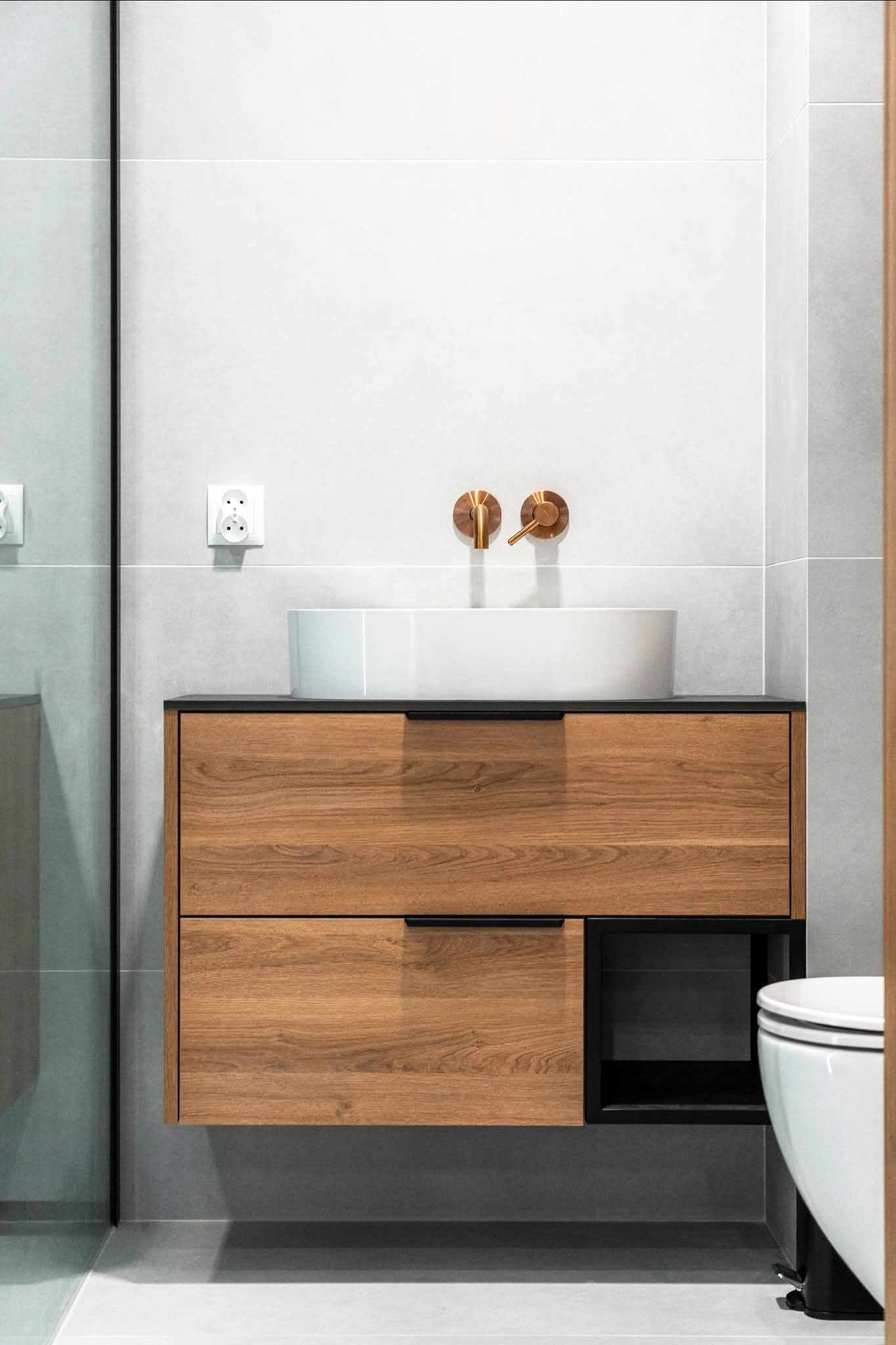 Nowoczesna łazienka: drewniana szafka pod umywalkę z czarnymi uchwytami i otwartą półką, okrągła umywalka nablatowa, miedziana armatura na tle szarych płytek.