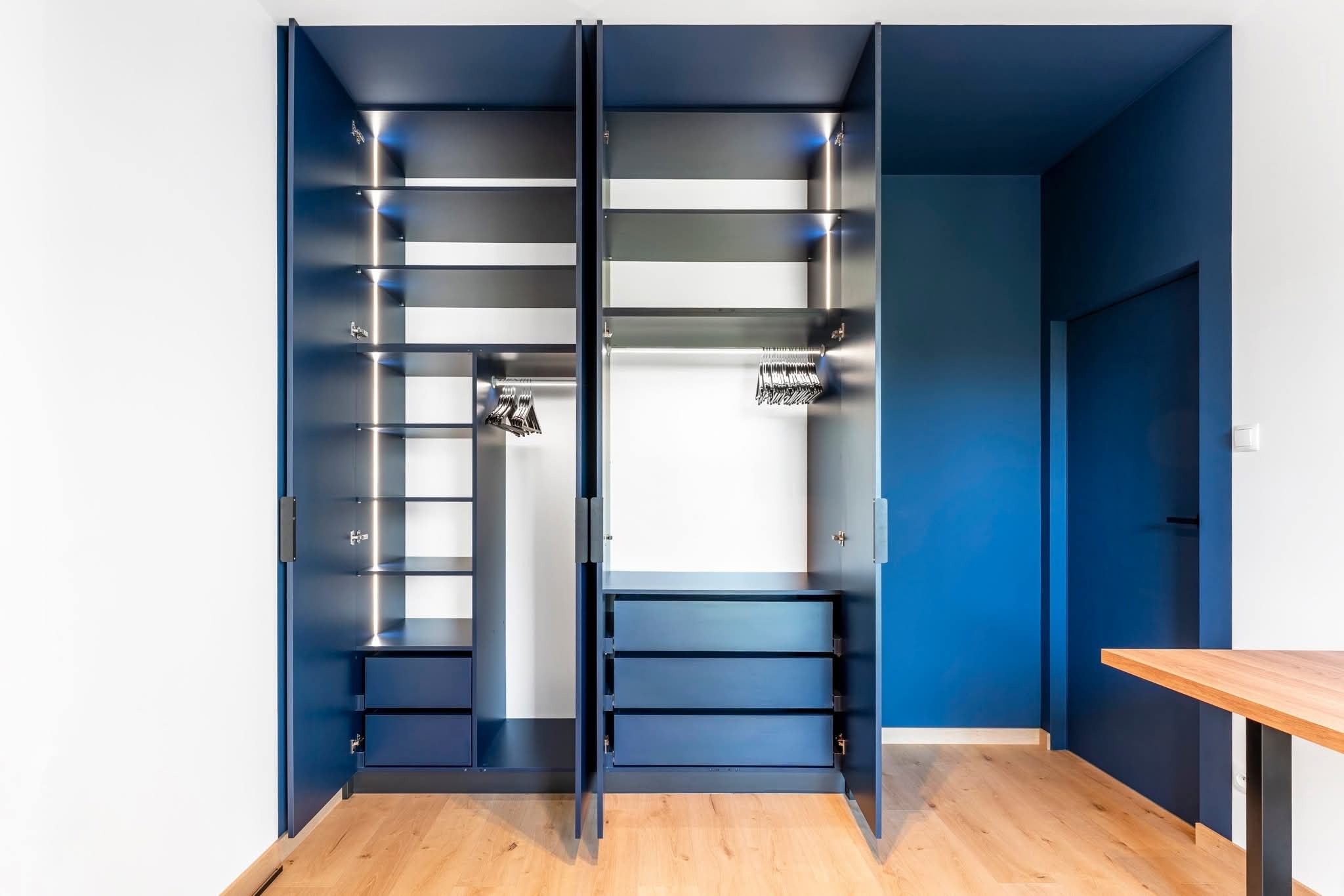 Niebieska szafa wnękowa z oświetleniem LED, otwarte drzwi ukazują półki, szuflady i wieszaki. Minimalistyczny design, drewniana podłoga, ściany białe i niebieskie.