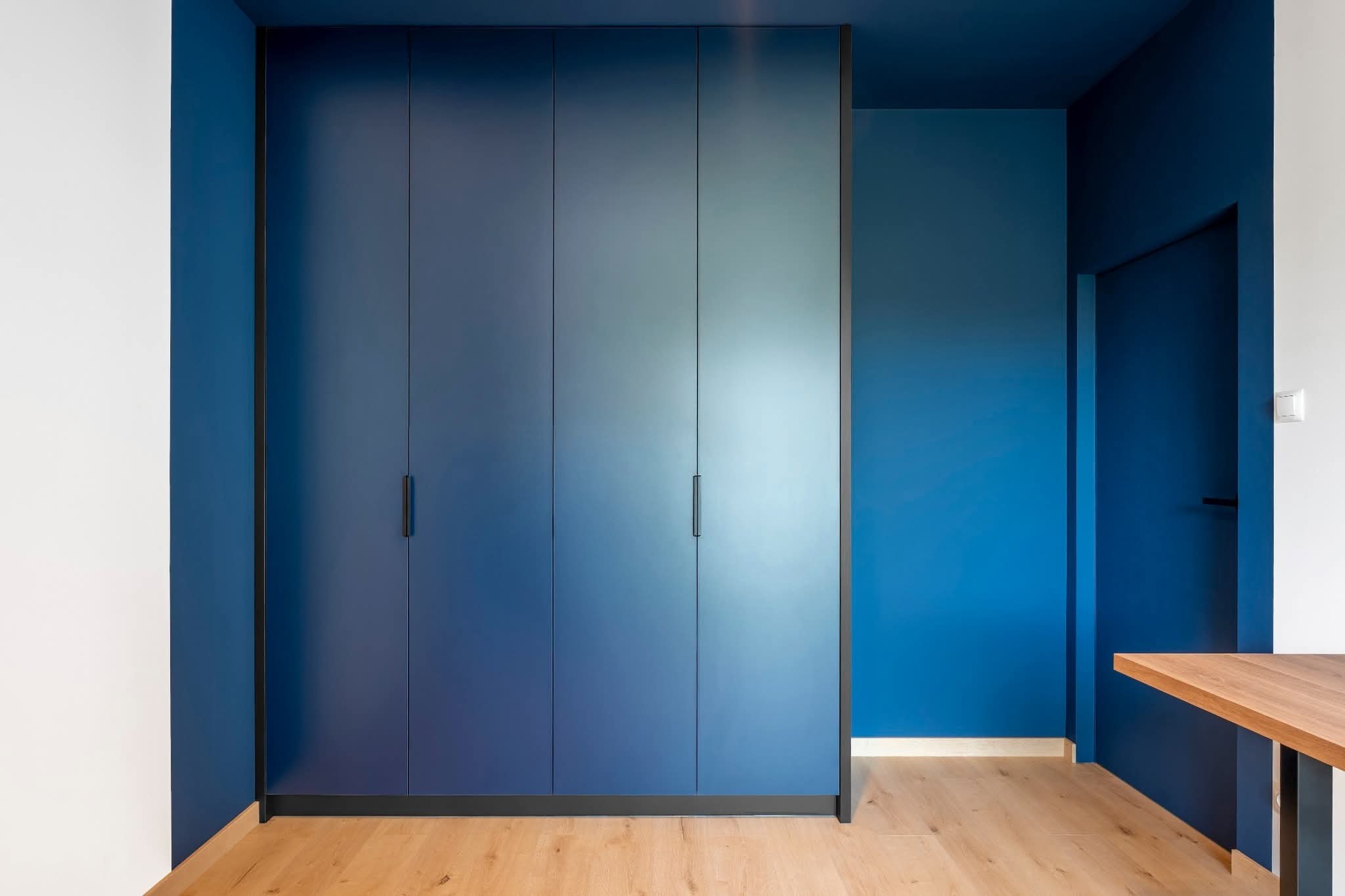 Niebieska szafa wnękowa z długimi, czarnymi uchwytami, dopasowana kolorystycznie do ścian i sufitu, na tle jasnej drewnianej podłogi. Minimalistyczny design.