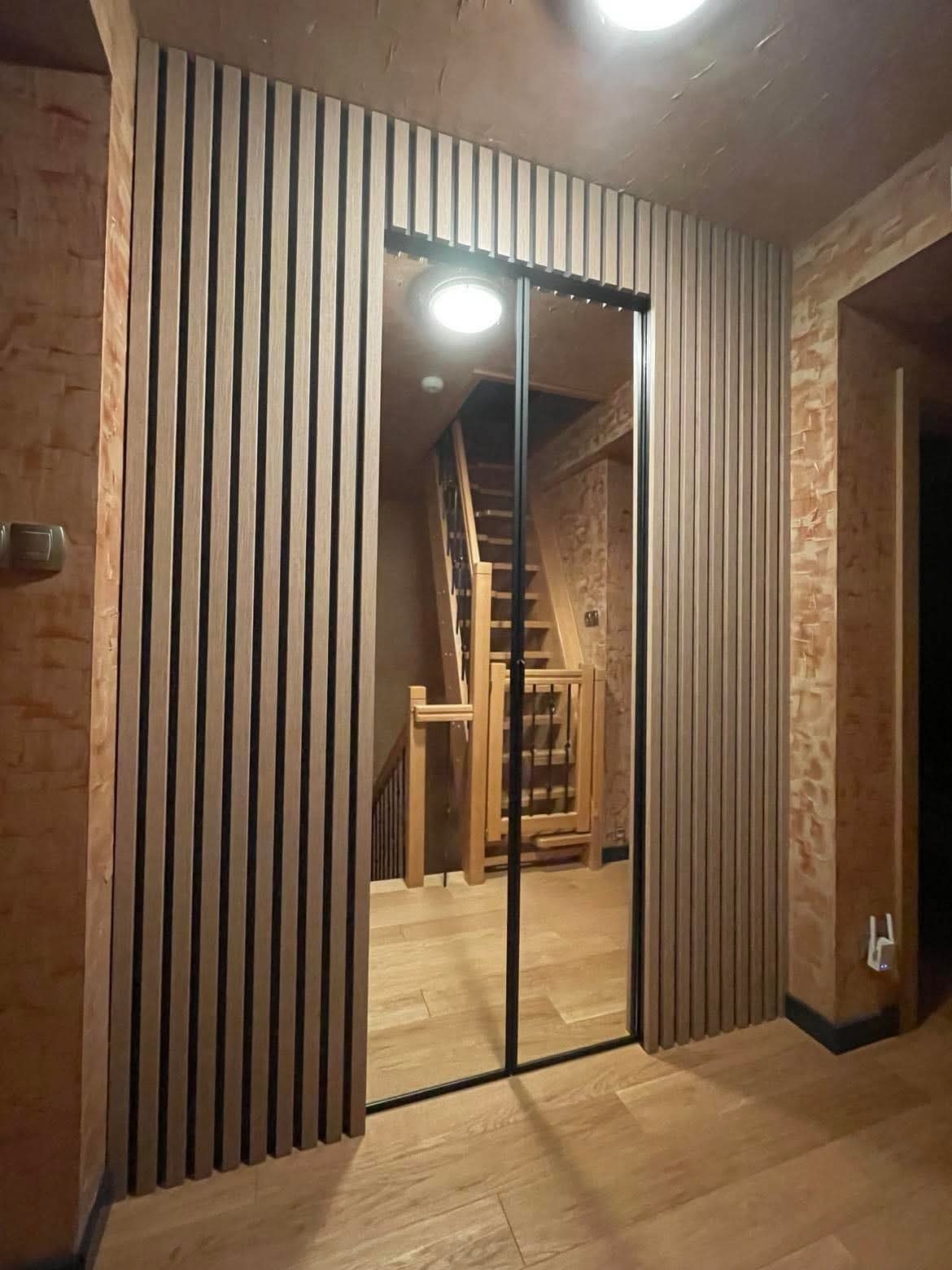 Nowoczesna szafa wnękowa z lustrzanymi drzwiami przesuwnymi i drewnianymi lamelami, wkomponowana w korytarz z drewnianymi schodami i podłogą.