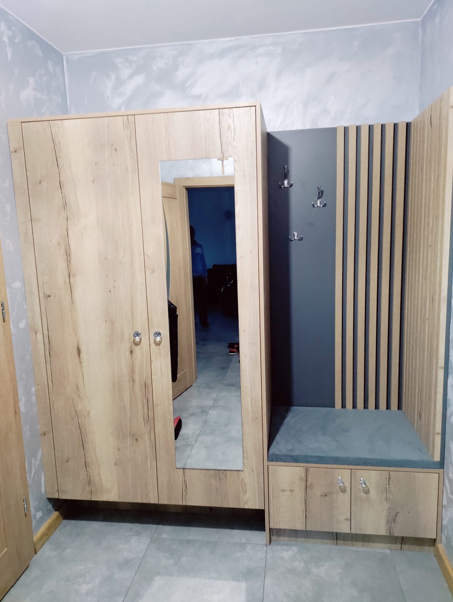 Komplet mebli do przedpokoju z lustrem i siedziskiem. Szafa z drewnopodobnymi frontami, szary panel z wieszakami i pionowe listwy dekoracyjne.