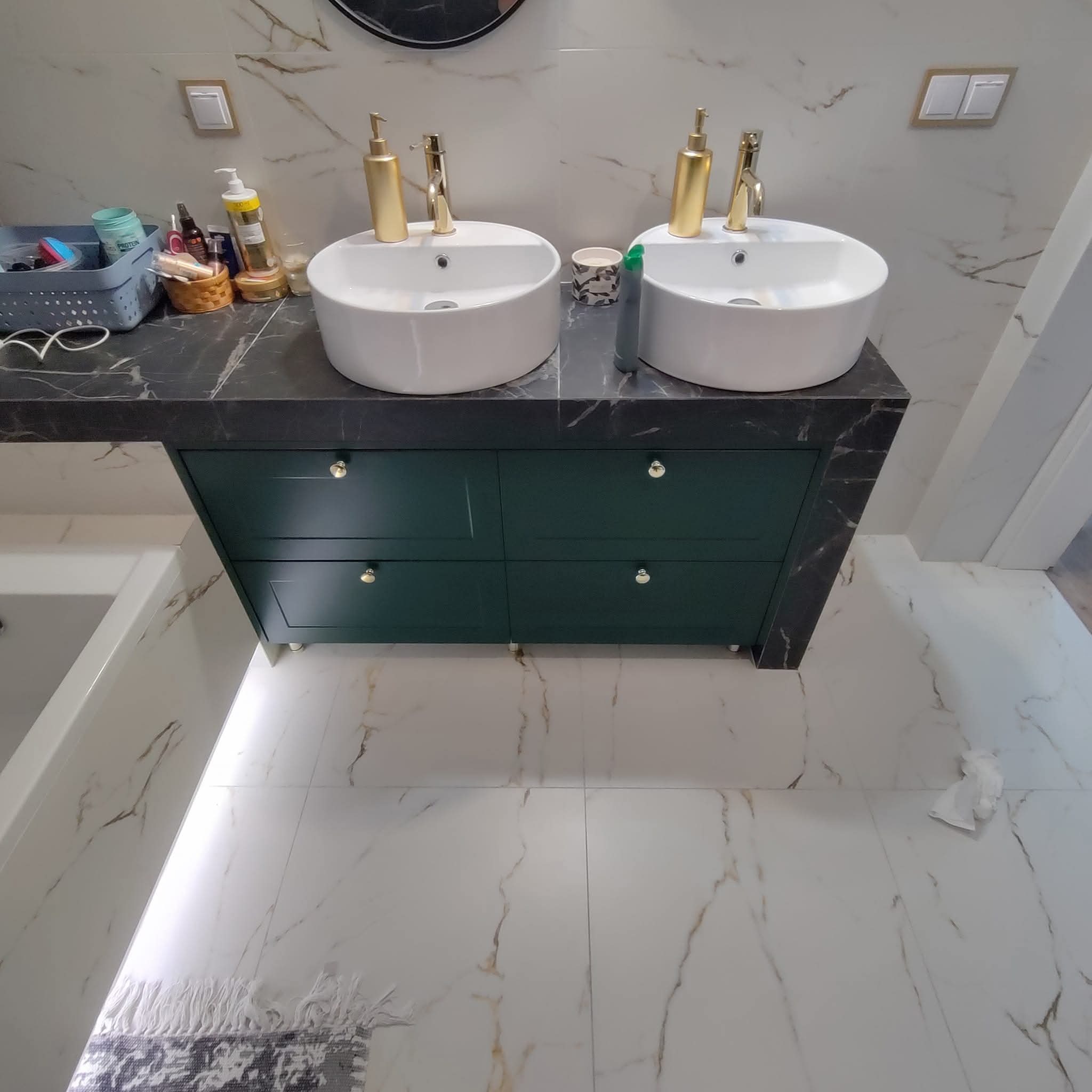 Nowoczesna łazienka z podwójną umywalką nablatową na ciemnozielonej szafce z blatem imitującym marmur. Białe płytki z marmurowym wzorem na ścianach i podłodze.