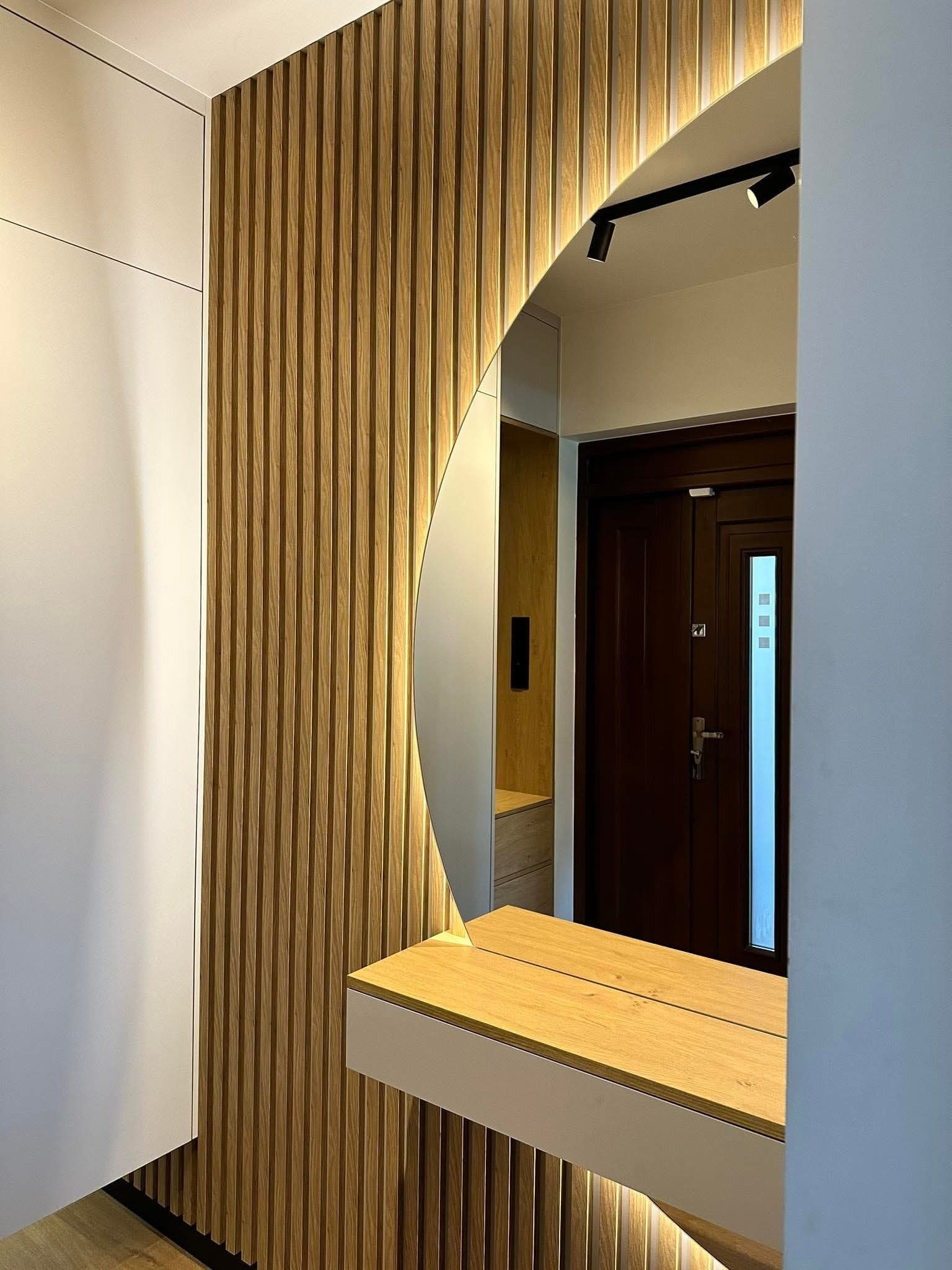 Nowoczesna aranżacja przedpokoju z okrągłym lustrem podświetlonym LED, drewnianą ścianą z lamelami i białą szafą w zabudowie. Widoczny fragment blatu z jasnego drewna.