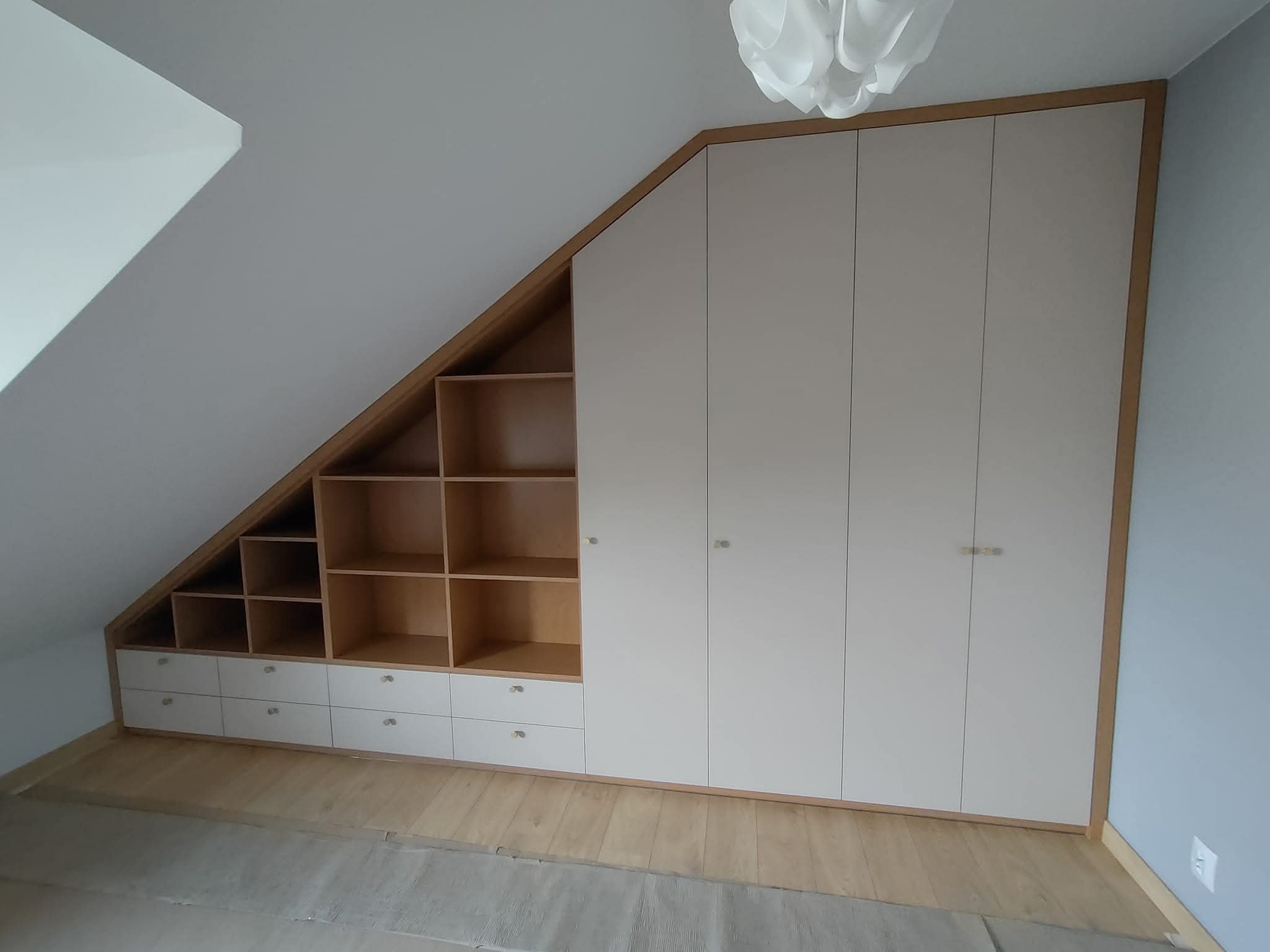 Szafa wnękowa na poddaszu ze skośnym sufitem, z otwartymi półkami i szufladami w dolnej części, oraz zamkniętymi szafkami po prawej stronie, wykończona drewnianą ramą.