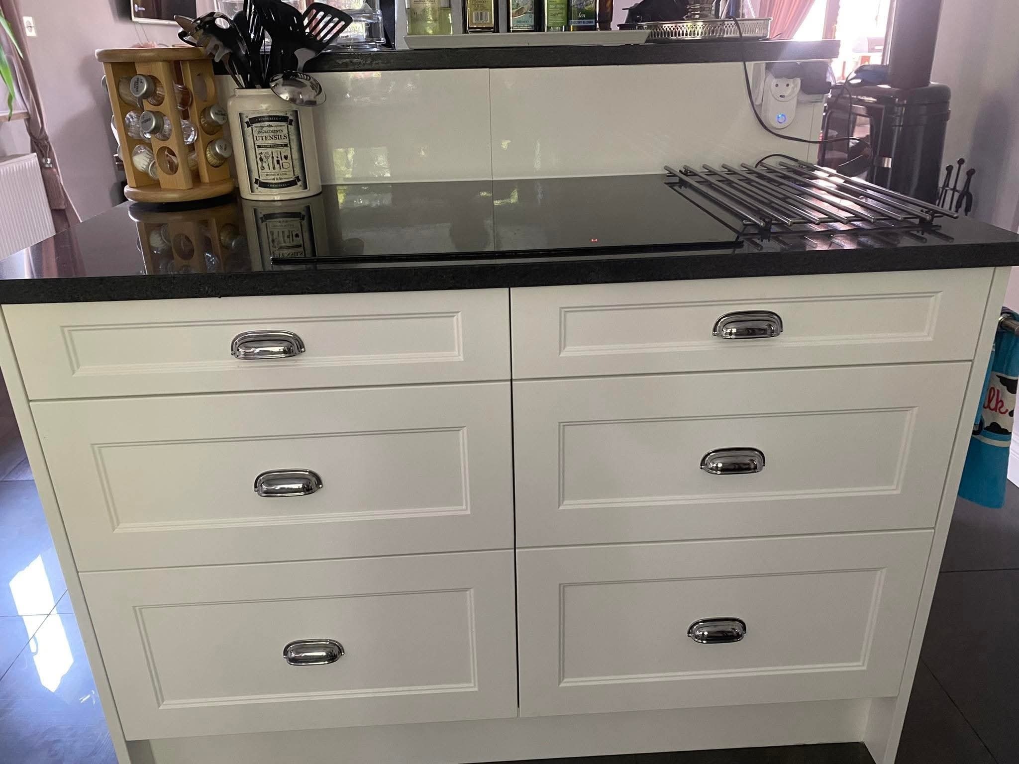 Białe szafki kuchenne z czarnym blatem, chromowane uchwyty, widoczne akcesoria kuchenne i płyta indukcyjna. Klasyczny styl.