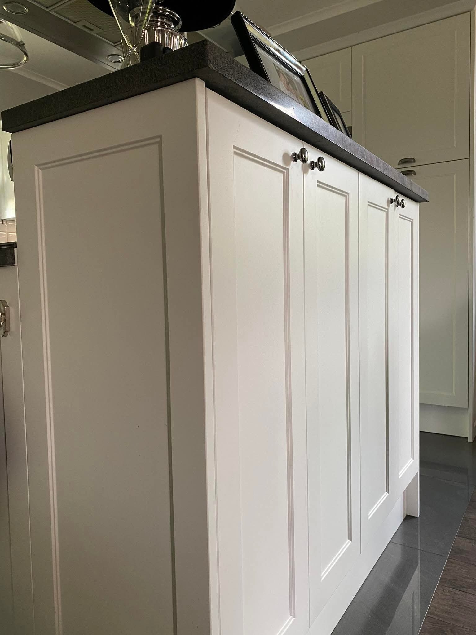 Biała wyspa kuchenna z ciemnym blatem, ozdobiona ramkami i gałkami. Na blacie dekoracje: ramki ze zdjęciami i szklane naczynia. Widoczne fragmenty zabudowy kuchennej w tle.