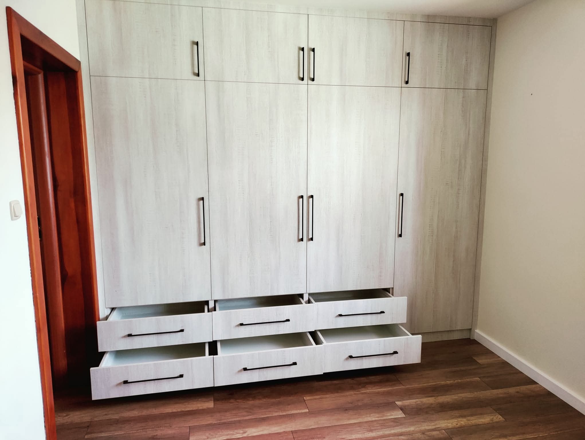 Jasna szafa wnękowa z szufladami i czarnymi uchwytami, na tle drewnianej podłogi. Część szuflad jest otwarta, ukazując białe wnętrze. Widoczny fragment brązowych drzwi.