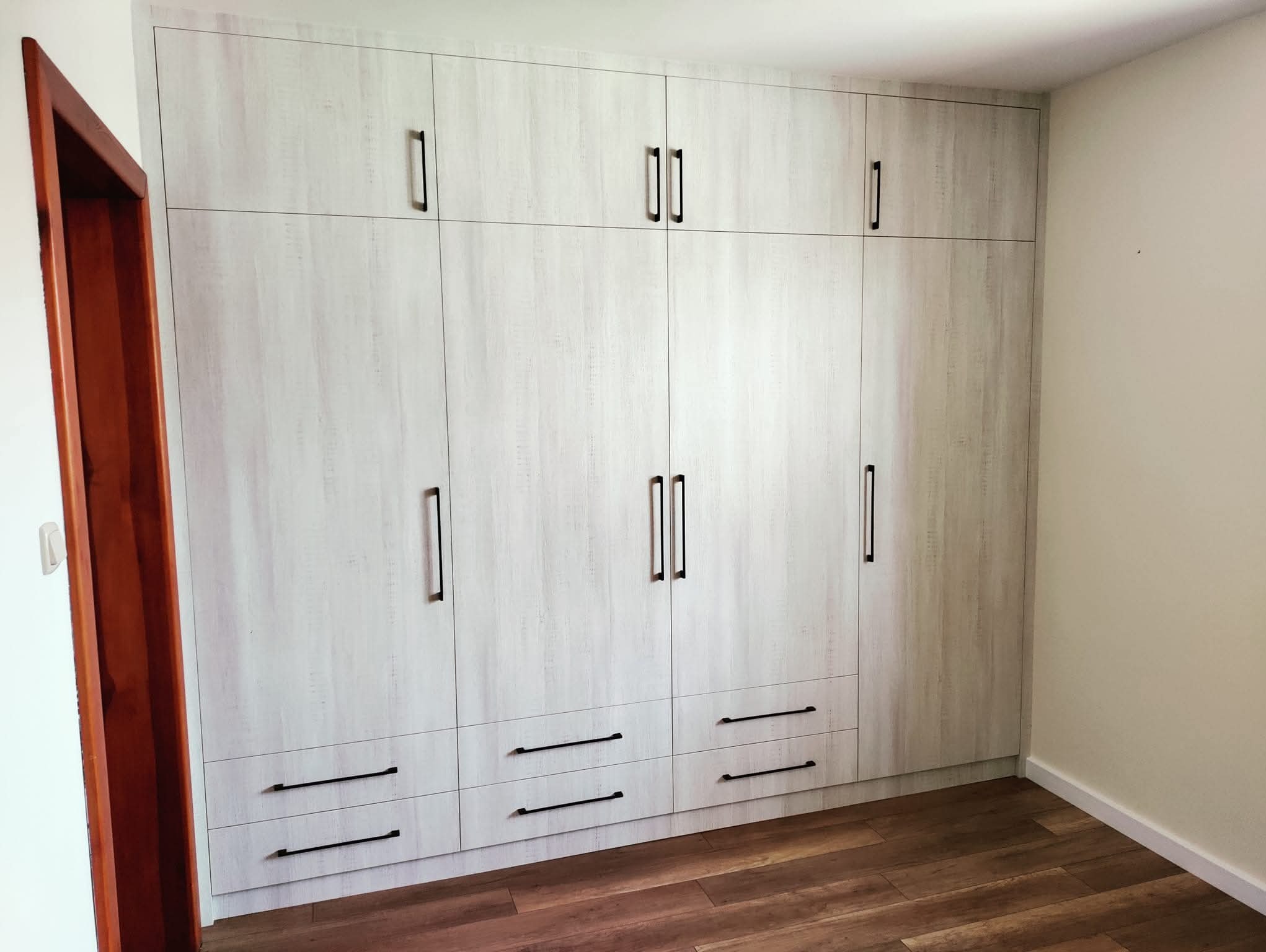 Jasna szafa wnękowa z drewnianymi uchwytami, dopasowana do podłogi. Widoczne drzwi wejściowe po lewej stronie. Minimalistyczny design, jasne wnętrze.