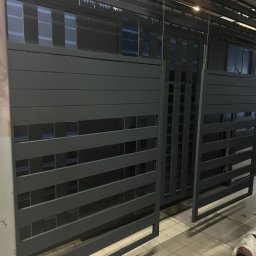 PHU Tomasz Dominikowski - Szare, metalowe ogrodzenie panelowe z poziomymi szczeblami, widoczne w hali produkcyjnej. Elementy czekają na montaż. Widoczne ślady po produkcji.