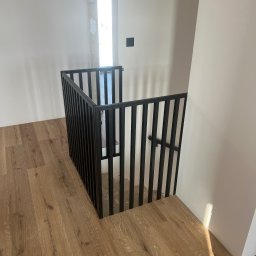 PHU Tomasz Dominikowski - Nowoczesna, czarna balustrada metalowa z pionowymi szczebelkami, zabezpieczająca schody w jasnym wnętrzu z drewnianą podłogą. Minimalistyczny design, surowe wykończenie.