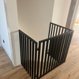 PHU Tomasz Dominikowski - Nowoczesna, czarna barierka schodowa z pionowych prętów, zamontowana w narożniku pomieszczenia z drewnianą podłogą. Minimalistyczny design, jasne ściany.