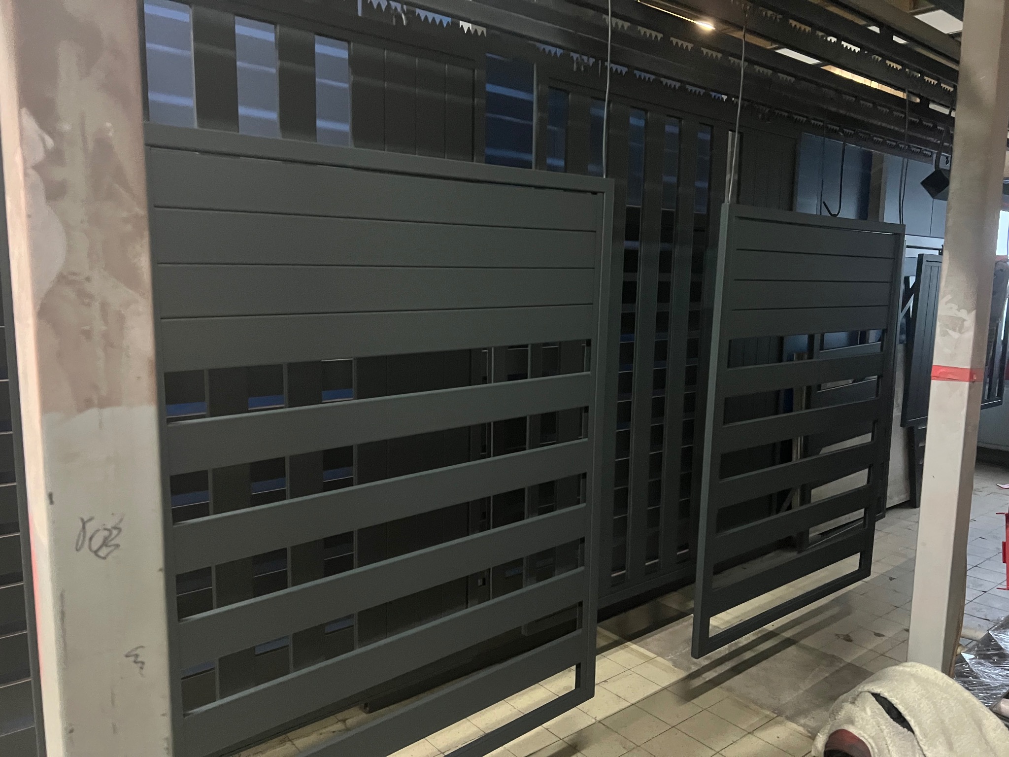 Szare, metalowe ogrodzenie panelowe z poziomymi szczeblami, widoczne w hali produkcyjnej. Elementy czekają na montaż. Widoczne ślady po produkcji.