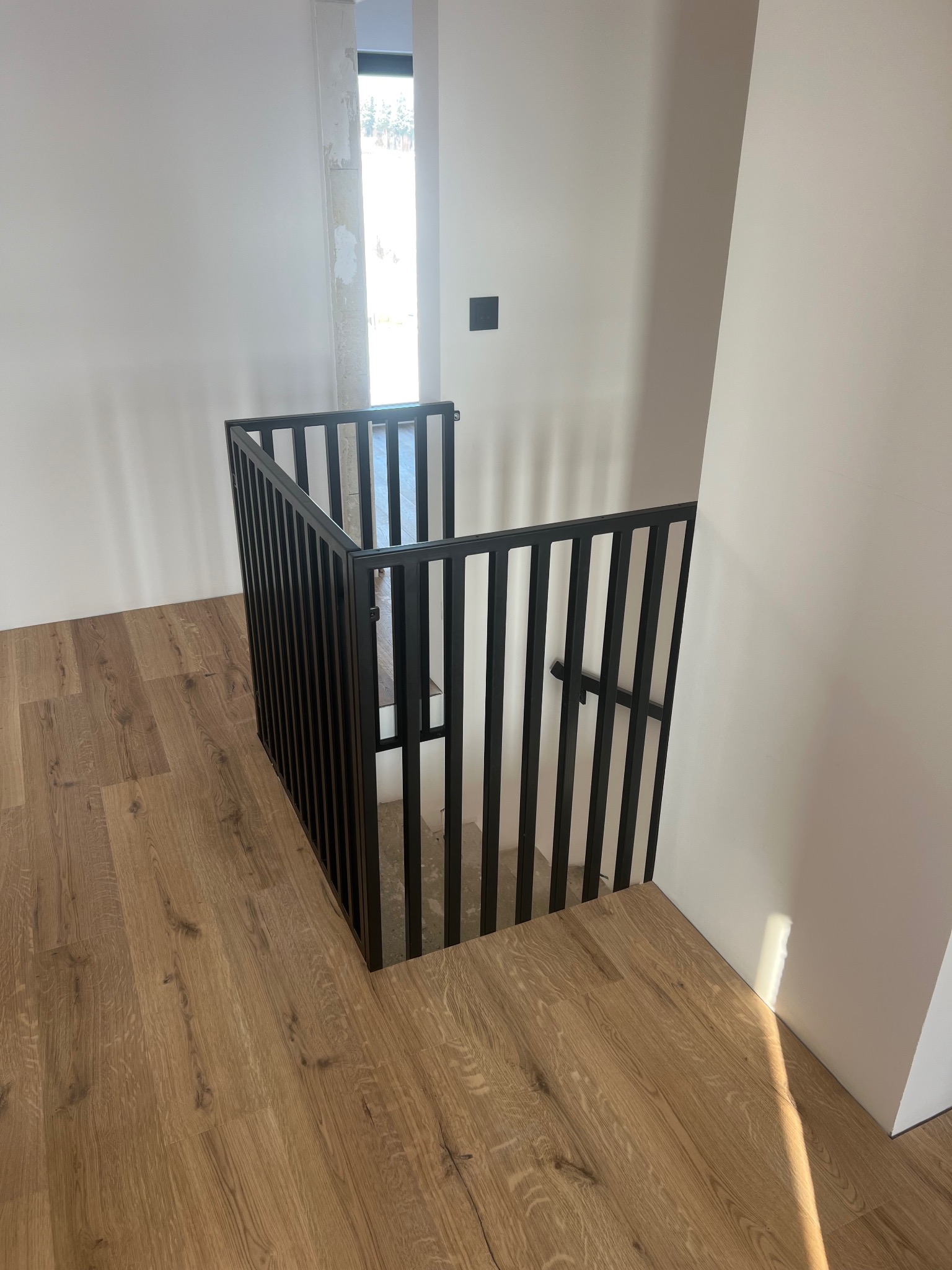 Nowoczesna, czarna balustrada metalowa z pionowymi szczebelkami, zabezpieczająca schody w jasnym wnętrzu z drewnianą podłogą. Minimalistyczny design, surowe wykończenie.