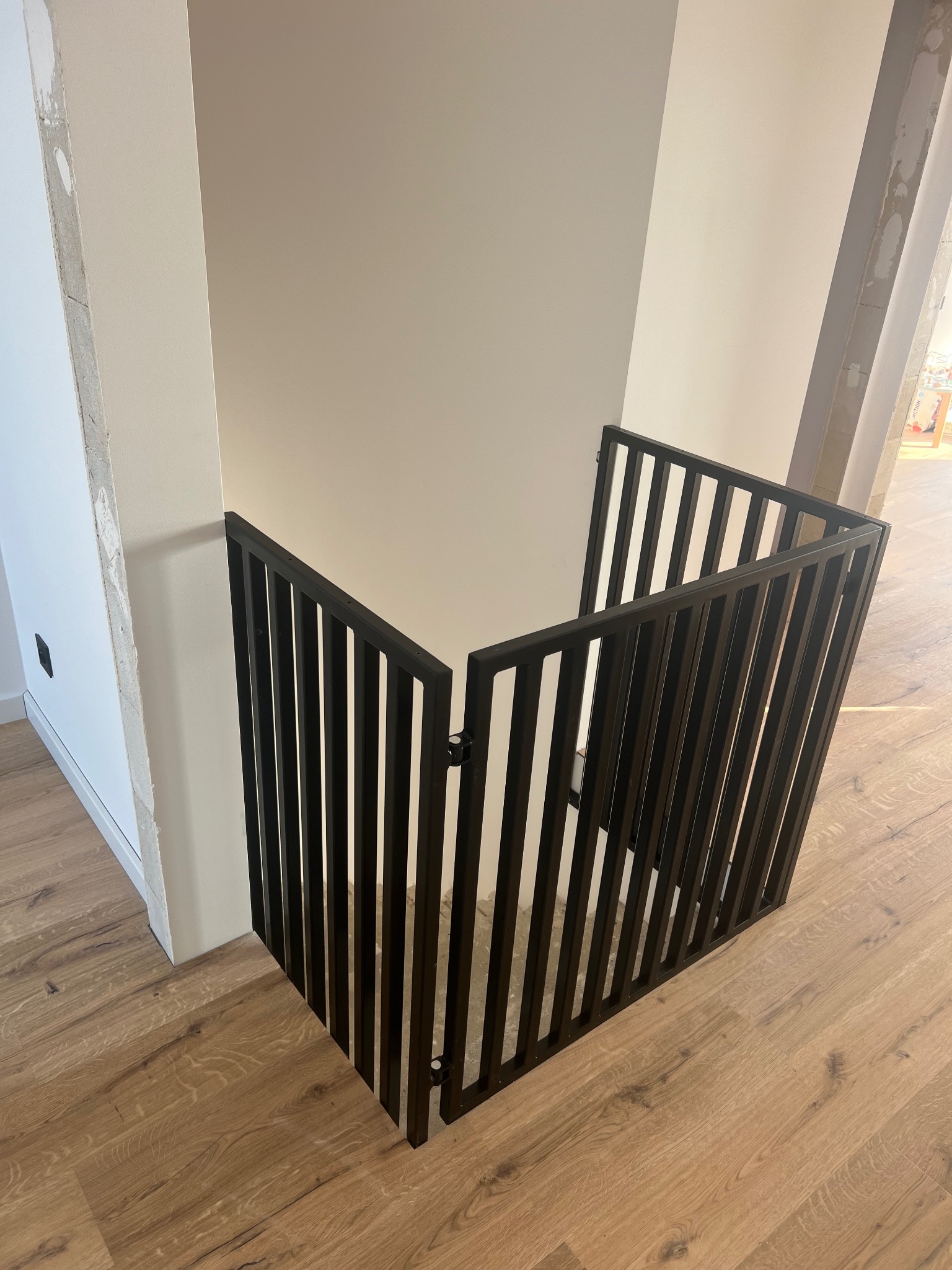 Nowoczesna, czarna barierka schodowa z pionowych prętów, zamontowana w narożniku pomieszczenia z drewnianą podłogą. Minimalistyczny design, jasne ściany.