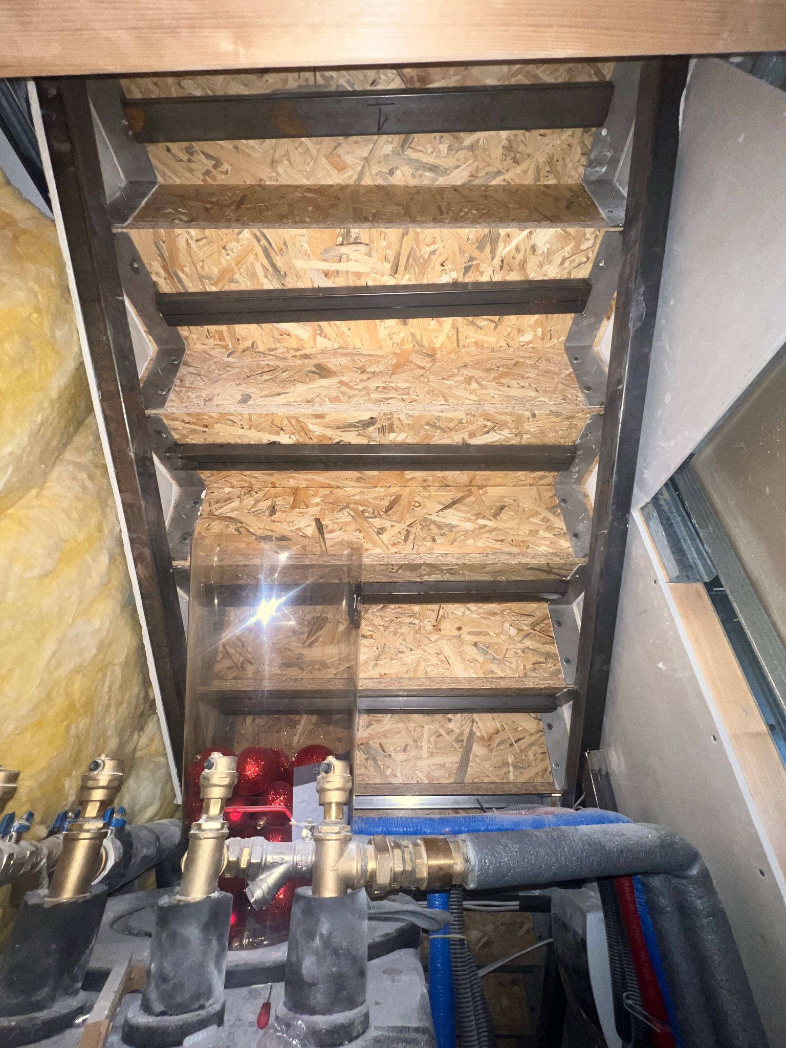 Metalowa konstrukcja schodów z płytami OSB, widok z dołu. W tle instalacja grzewcza i ocieplenie. Surowy stan, etap budowy.