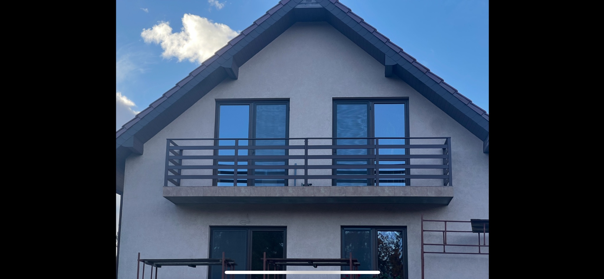 Dwukondygnacyjny dom z balkonem zabezpieczonym nowoczesną, poziomą balustradą w kolorze grafitowym. Błękitne niebo w tle. Częściowo widoczne rusztowanie.