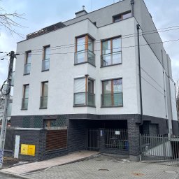 Nowoczesny, trzypiętrowy budynek mieszkalny z garażem w przyziemiu, wykończony w bieli i szarości, z drewnianymi ramami okiennymi i balkonami z balustradami.