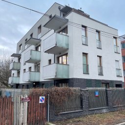 Nowoczesny, trzypiętrowy budynek mieszkalny z balkonami, wykończony w bieli i szarości, z elementami drewnianymi. Widok z poziomu ulicy, pochmurne niebo.