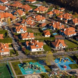 Osiedle domów jednorodzinnych z czerwonymi dachami, widok z lotu ptaka, z placem zabaw i boiskiem w kadrze. Jesienne drzewa w tle.