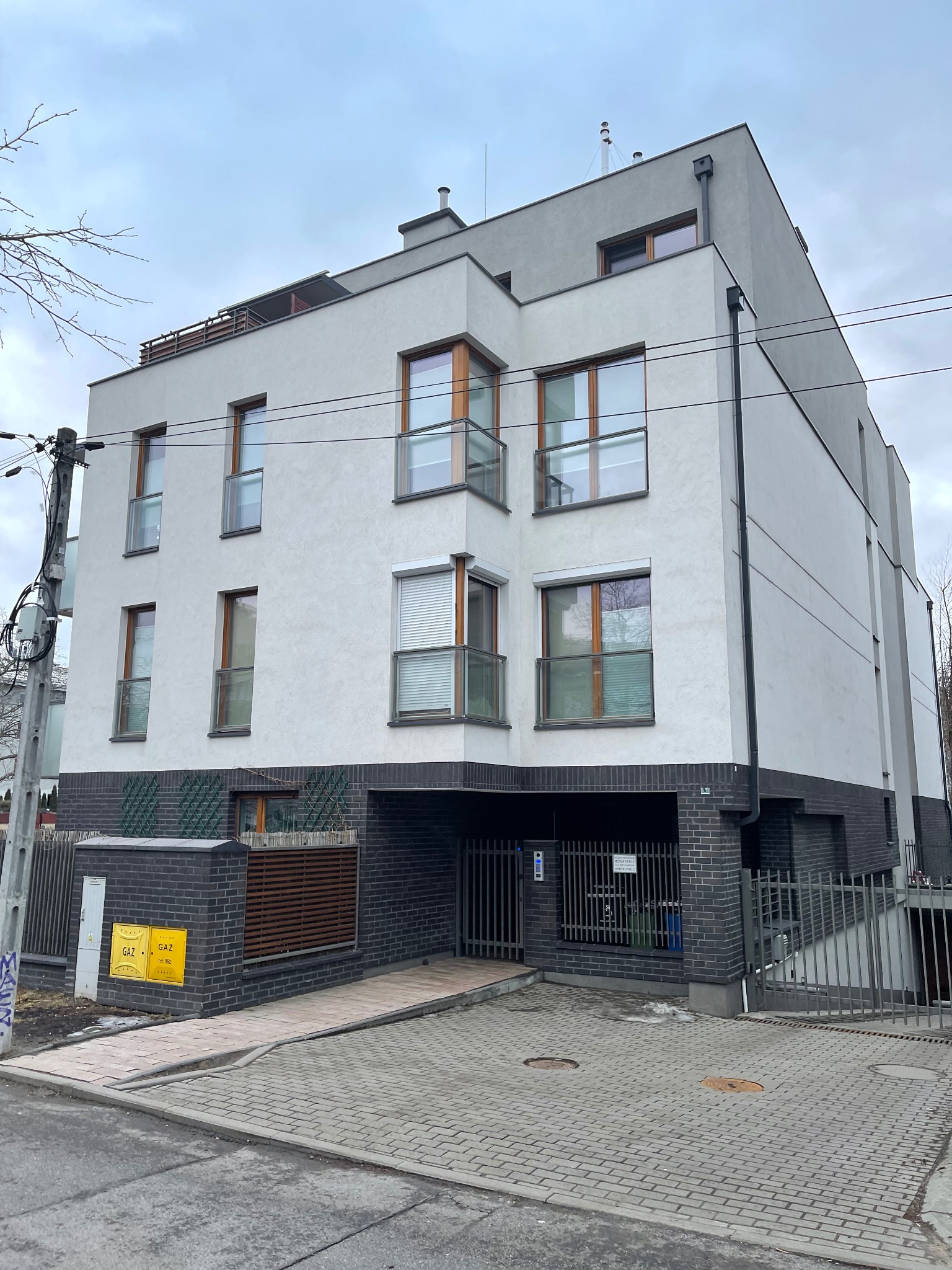 Nowoczesny, trzypiętrowy budynek mieszkalny z garażem w przyziemiu, wykończony w bieli i szarości, z drewnianymi ramami okiennymi i balkonami z balustradami.