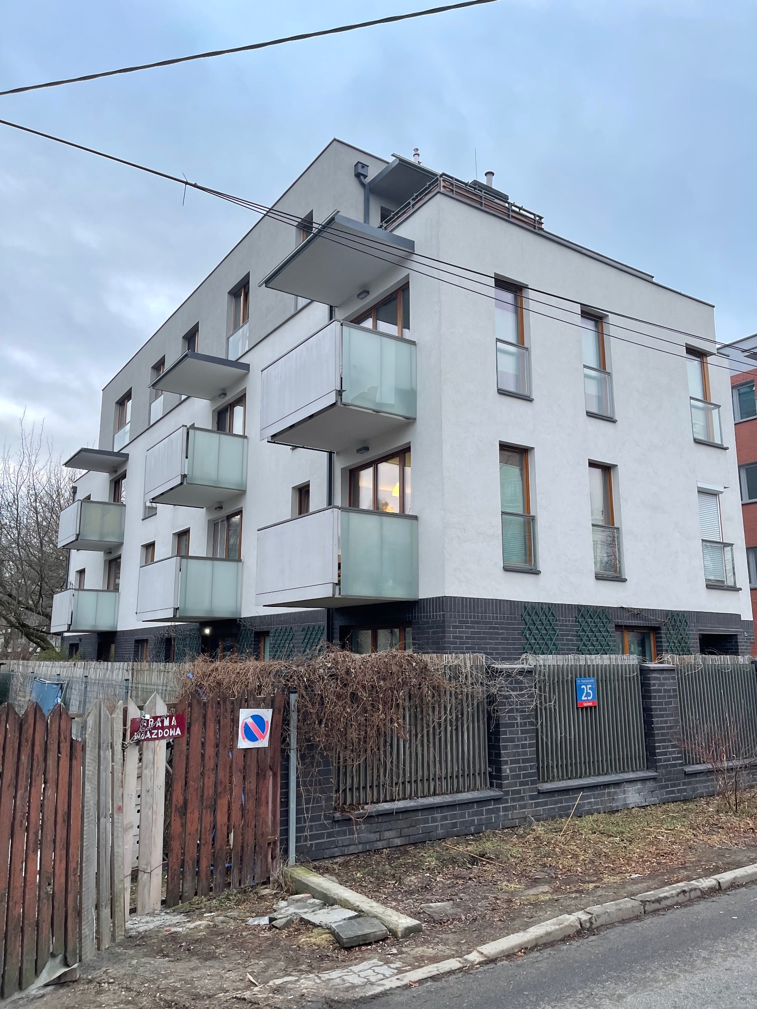 Nowoczesny, trzypiętrowy budynek mieszkalny z balkonami, wykończony w bieli i szarości, z elementami drewnianymi. Widok z poziomu ulicy, pochmurne niebo.