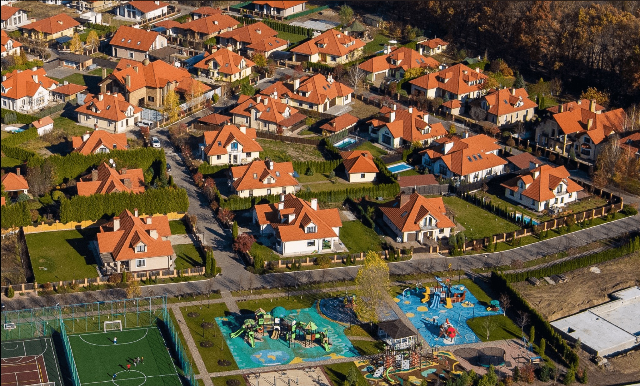 Osiedle domów jednorodzinnych z czerwonymi dachami, widok z lotu ptaka, z placem zabaw i boiskiem w kadrze. Jesienne drzewa w tle.