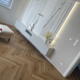 Michał Pawelec - Eleganckie wykończenie wnętrza: parkiet w jodełkę, ściana z imitacją marmuru, biała zabudowa meblowa z dekoracjami i subtelnym oświetleniem.