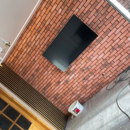 Michał Pawelec - Ściana z cegły klinkierowej z wnęką na TV i panelami dekoracyjnymi w Turobinie. Widoczny fragment drzwi i wiadro z farbą. Nietypowy kąt ujęcia.