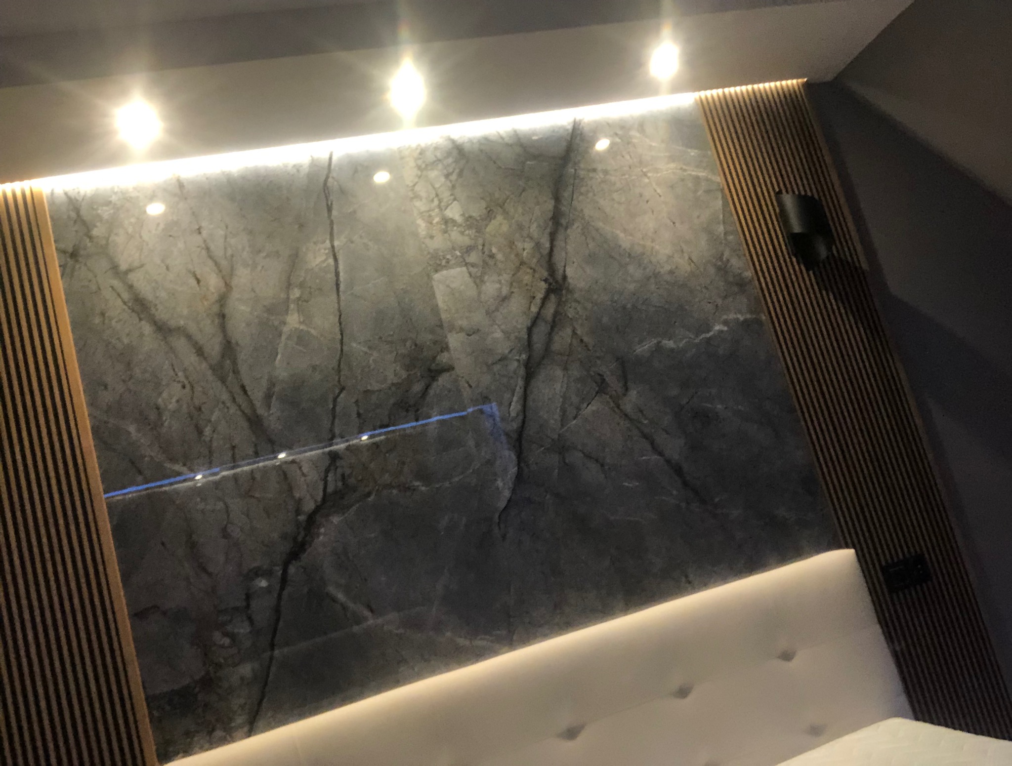 Ściana z szarego marmuru z drewnianymi panelami i oświetleniem LED nad wezgłowiem łóżka. Nowoczesny design wnętrza sypialni z akcentem na naturalne materiały.