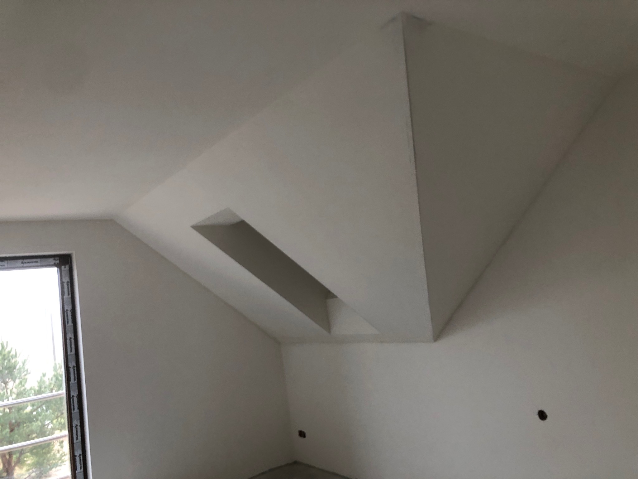 Białe, świeżo pomalowane poddasze z oknem i nietypową konstrukcją sufitu z wnęką. Widoczne ślady po szpachlowaniu i przygotowaniu do dalszych prac wykończeniowych.