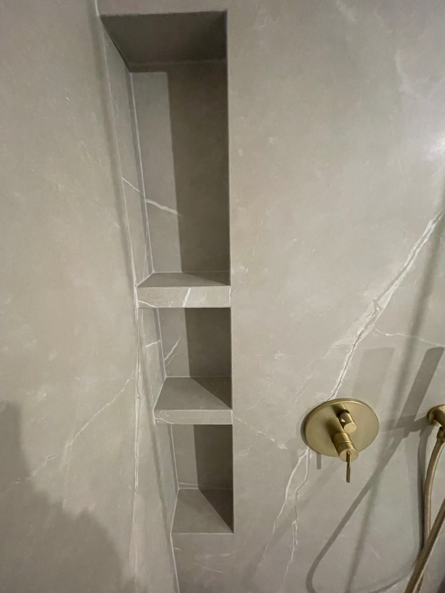 Wykończona wnęka z półkami w łazience, obłożona płytkami imitującymi marmur. Złota bateria natryskowa. Minimalistyczny design.