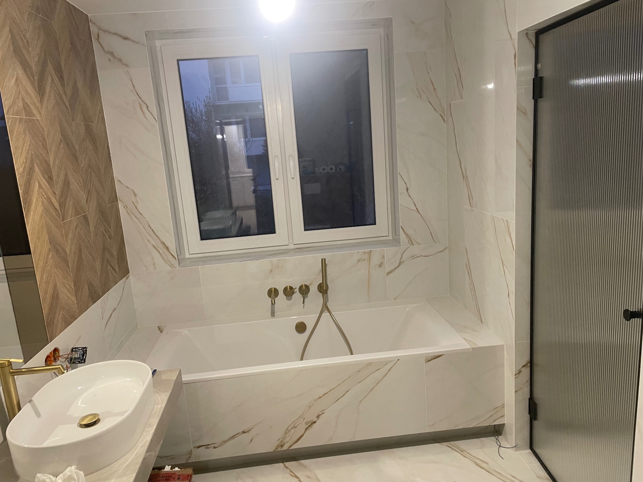 Elegancka łazienka w bieli i złocie: marmurowe płytki, wolnostojąca wanna z złotą armaturą, umywalka nablatowa i kabina prysznicowa z ryflowanego szkła.